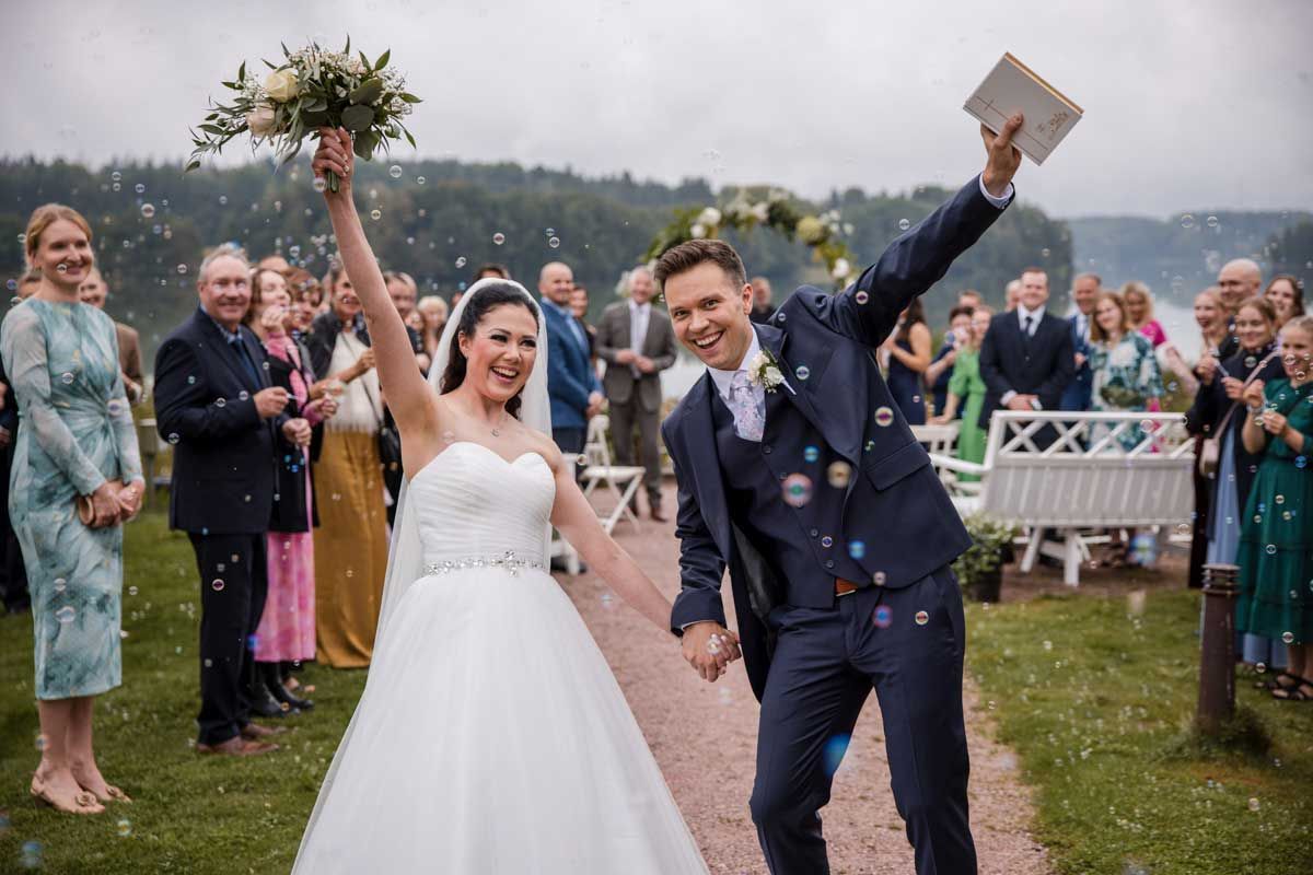 Hääpari heti vihkimisen jälkeen. Vieraat puhaltaa saippuakuplia. Hääpari tuulettaa kameralle, morsiammella kimppu ja sulhasella raamattu.