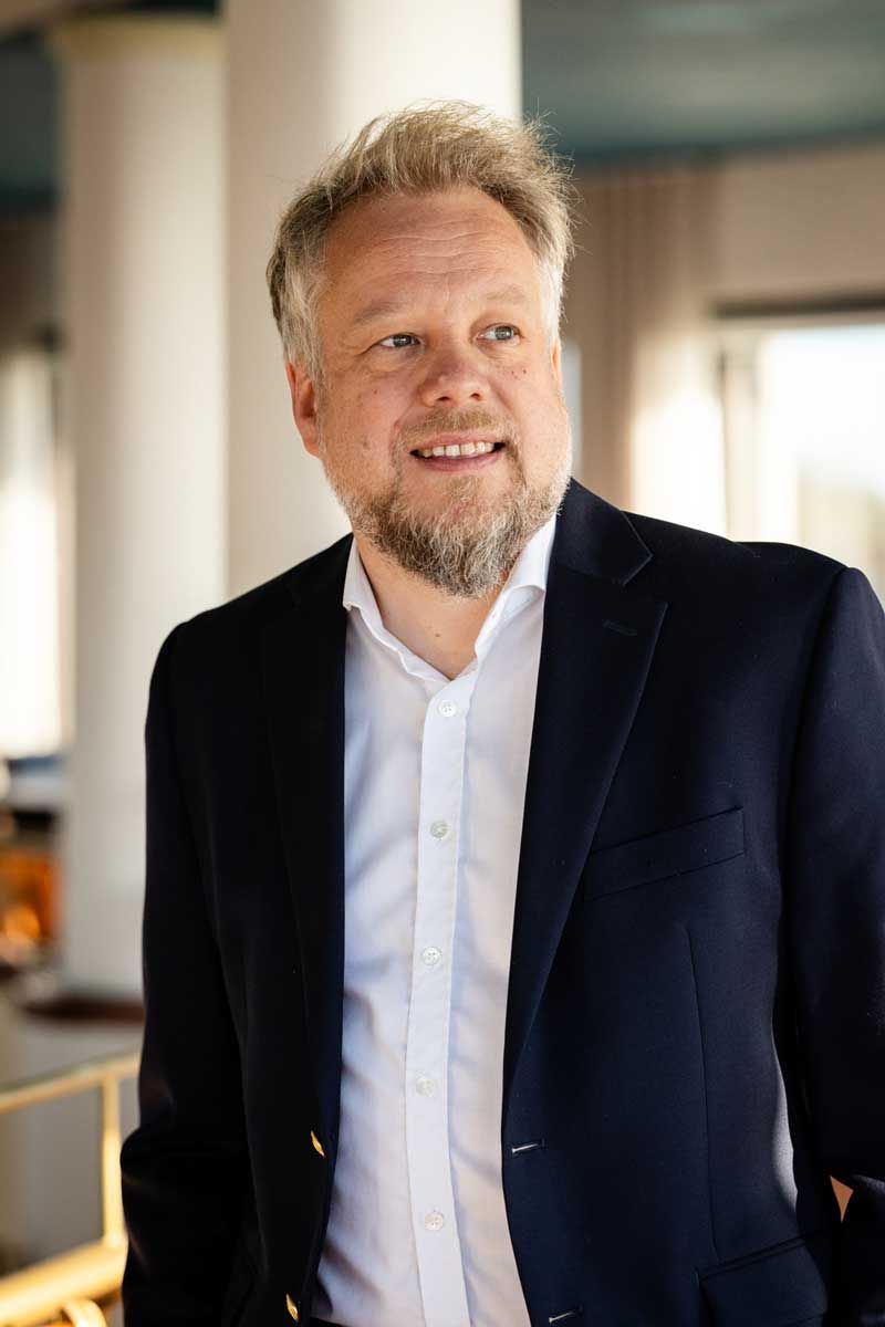 Keski-ikäinen mies puvun takissa valkoinen kauluspaita päällä. Rintakuva, katse ohi kameran ja luonnollinen ilme. Kuva otettu kahvilassa.
