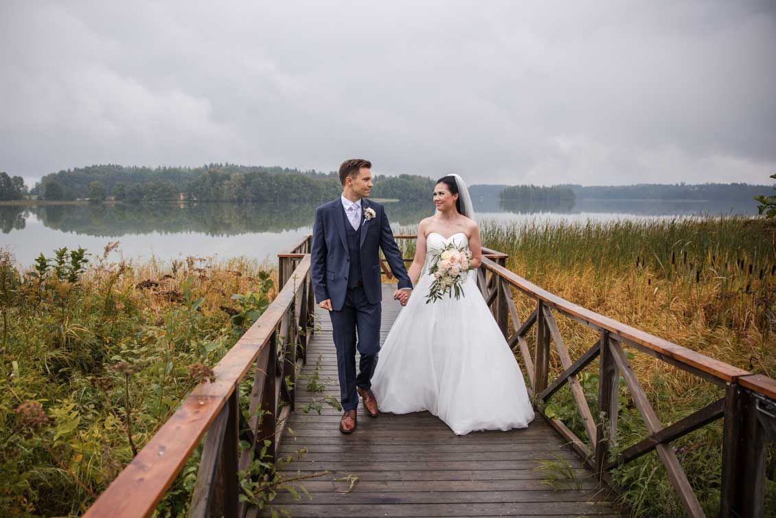Sulhanen ja morsian kävelee käsi kädessä laiturilla sumuisena ja harmaana päivänä. Morsiammella hääkimppu kädessä. Järvi taustalla.