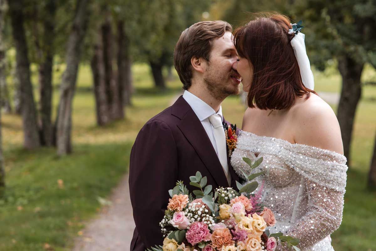 Morsian ja sulhanen on juuri suutelemassa ulkona tammikujalla. Morsiammella on kimalteleva hääpuku päällä ja hääkimppu kädessä.