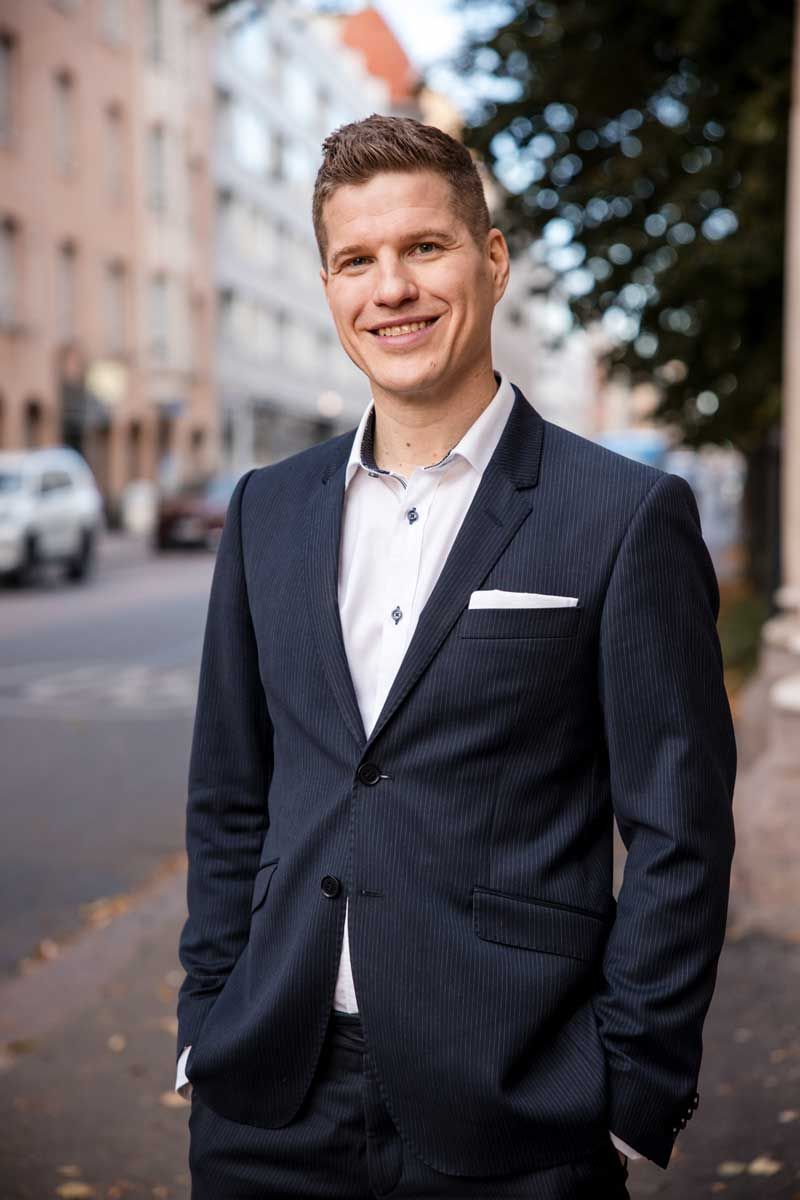Komea mies seisoo kadulla Helsingissä tummansinen puvun takki päällä. Katsoo rennosti kameraan ja hymyilee.