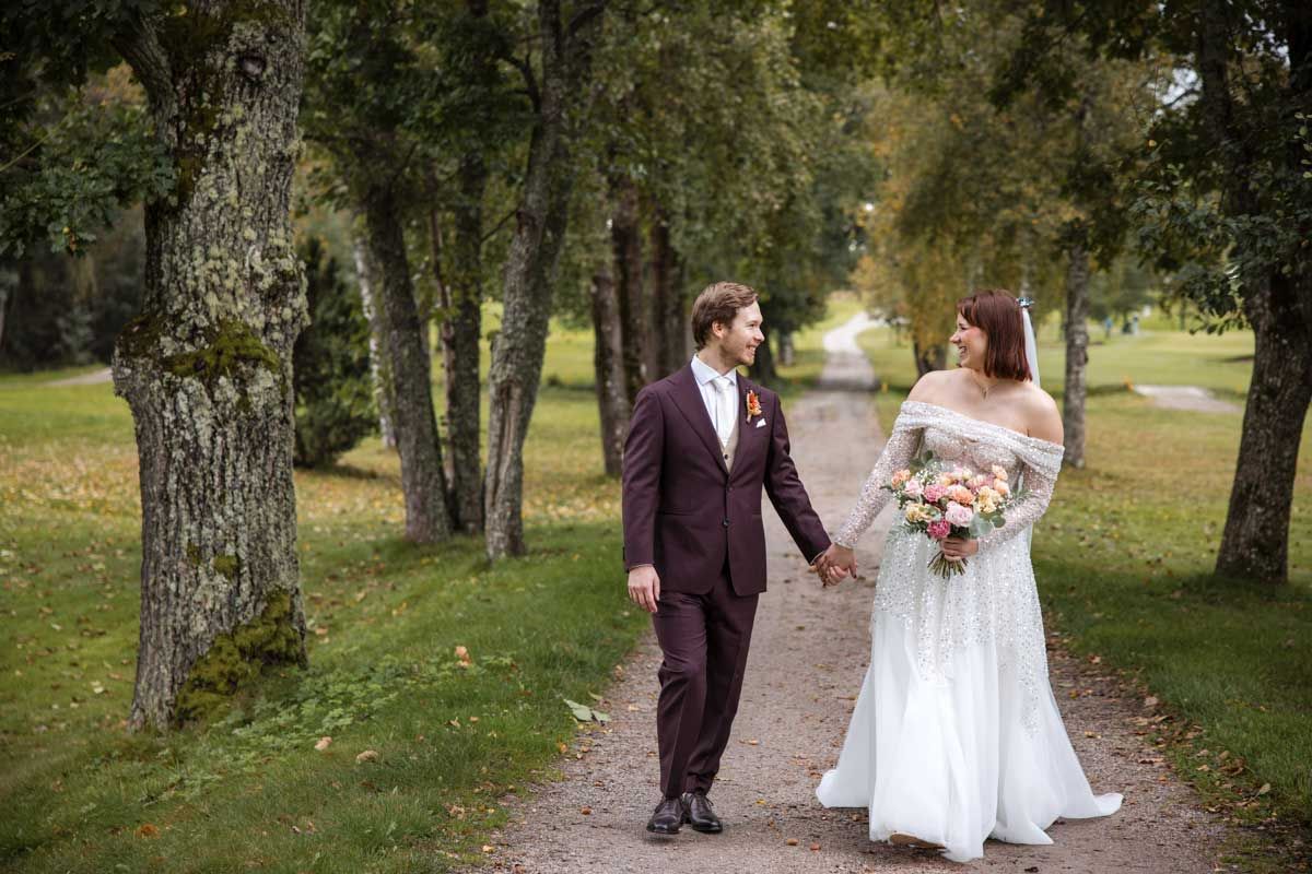 Hääpari kävelee tiellä mitä reunustaa tammet. Hääpari pitää käsistä kiinni ja katsoo toisiaan. Morsiammella on hääkimppu kädessä.