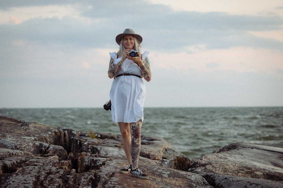 Valokuvaaja Suvi-Tuuli Kiilunen seisoo kalliolla meren äärellä. Valkoinen mekko päällä, filmikamera kädessä. Harmaa hattu päässä.