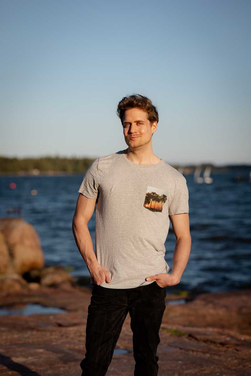 Nuori komea mies seisoo meren rannalla kalliolla. Harmaa t-paita päällä. Ilta-aurinko valaisee kasvot ja vartalon kauniisti. Meri taustalla.