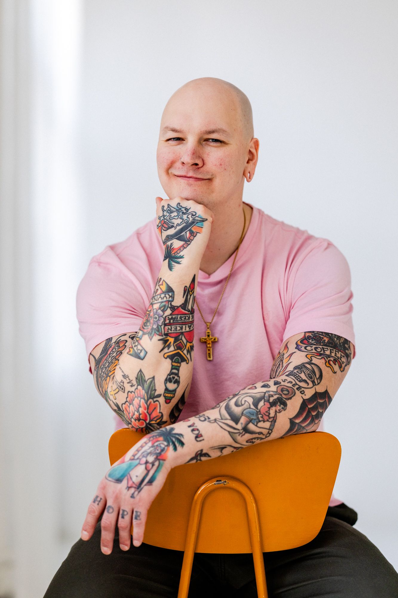 Kalju tatuoitu mies pinkissä t-paidassa. Mies istuu tuolilla ja pitää käsiä tuolin selkänojaa vasten. Mies katsoo suoraan kameraan.