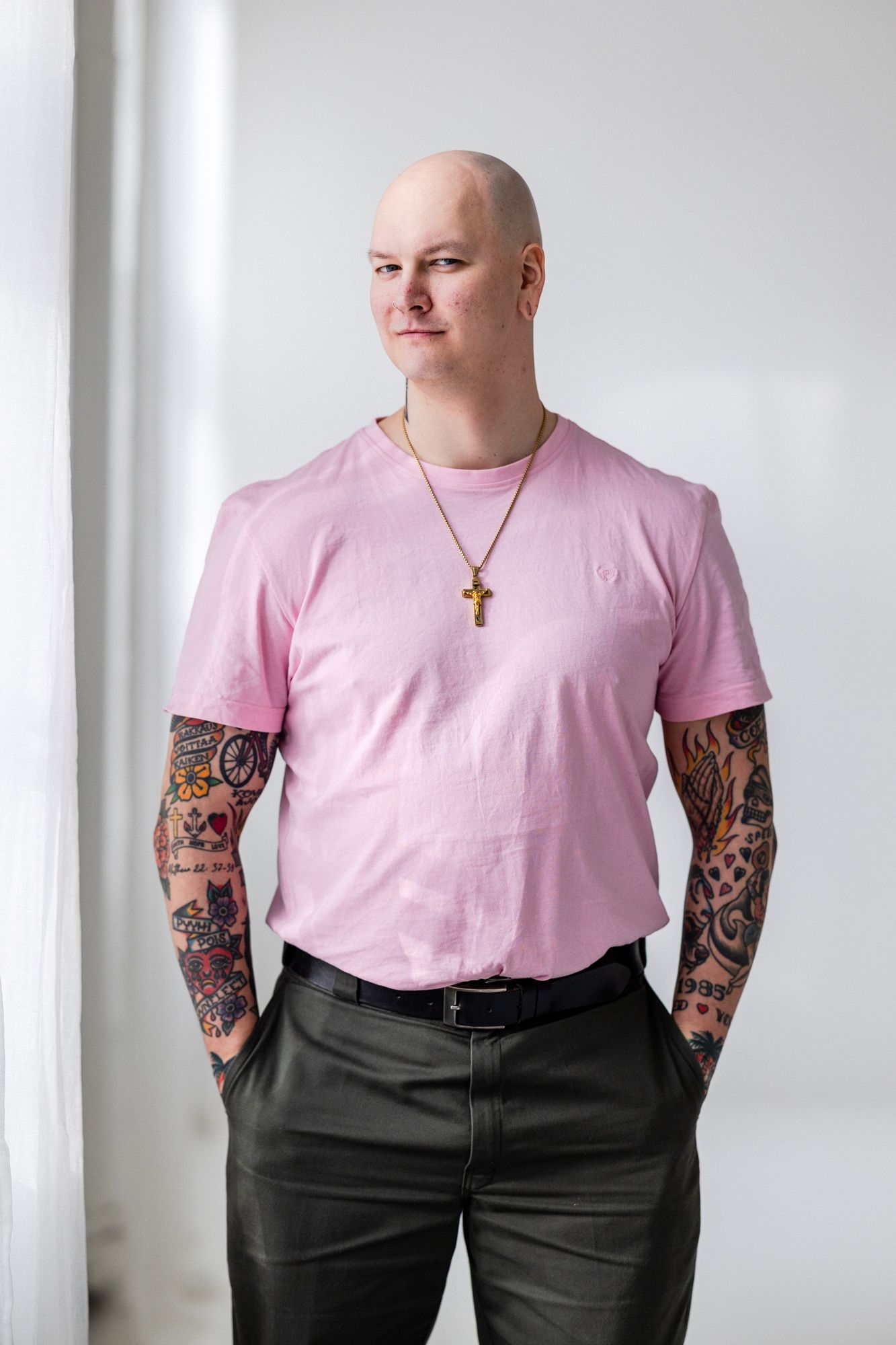 Tatuoitu kalju mies seisoo. Miehellä on pinkki t-paita päällä ja iso risti kaulassa. Mies seisoo vaaleassa studiotilassa.