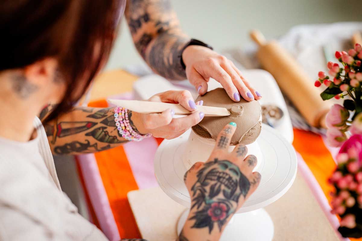 Ohjaajan ja asiakkaan tatuoidut kädet työstää keramiikkaa. Ohjaaja näyttää miten saviastiaan tehdään jalka. Oppilas seuraa vierestä.