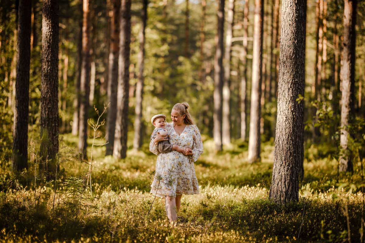 Äiti kävelee tunnelmallisessa metsässä taapero sylissä kesäisen kauniissa iltavalossa. Äiti katsoo vauvaa, vauva kurkistaa kameraan.