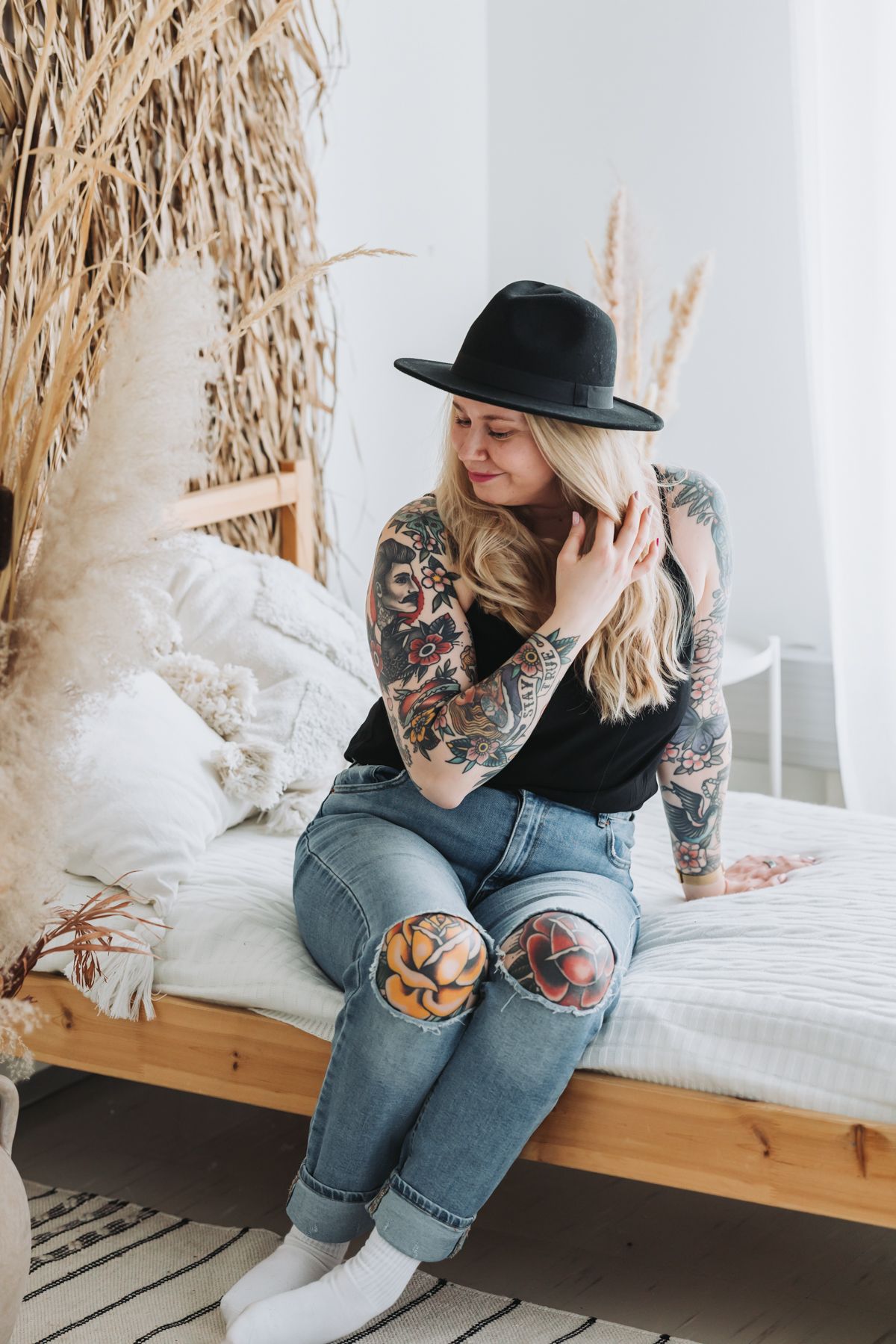 Valokuvaaja Suvi-Tuuli Kiilunen istuu sängyllä farkut ja musta toppi päällä. Tatuoidut kädet näkyvillä haroo hiuksiaan hattu päässä.