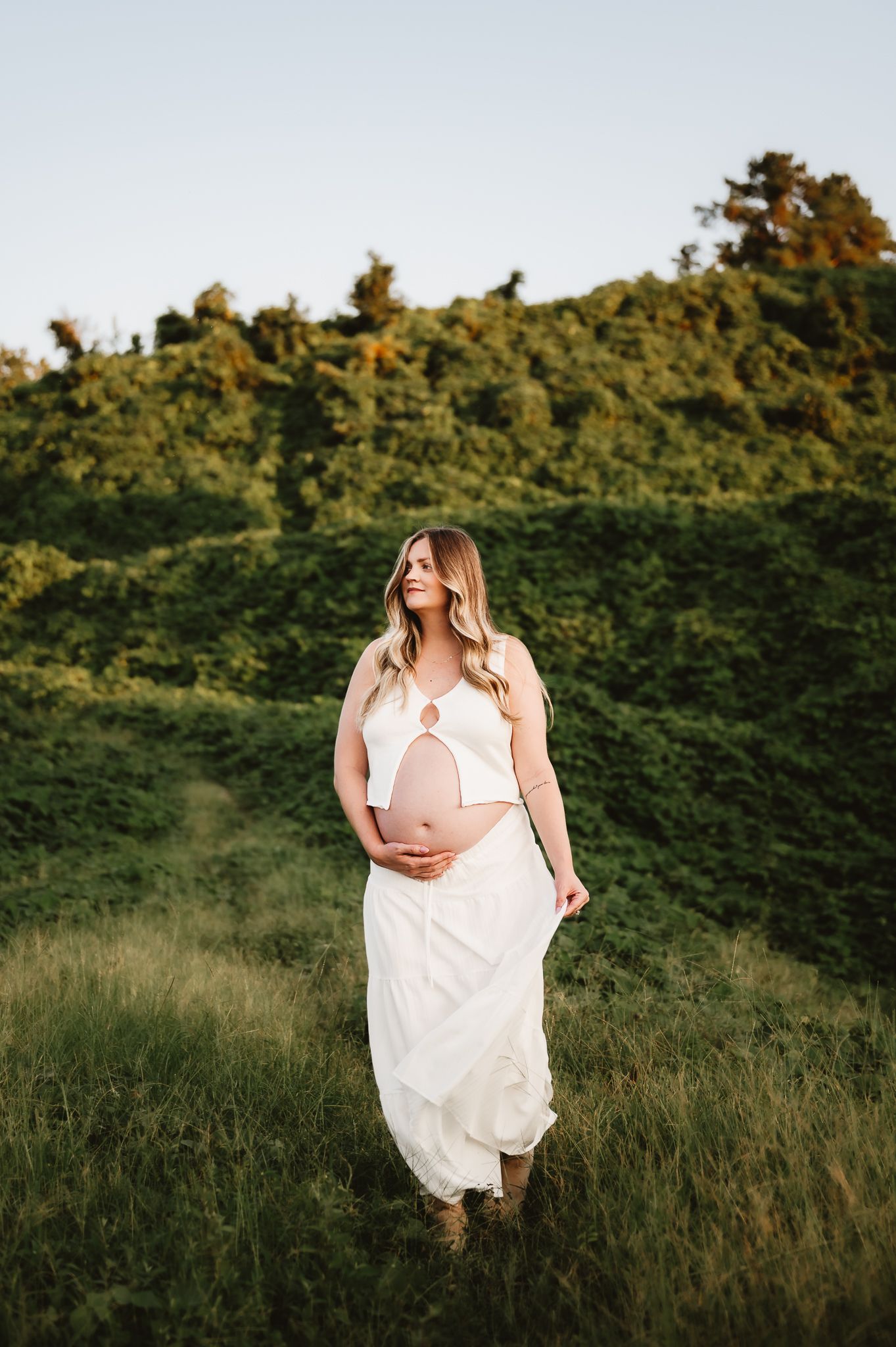 A figure in a flowing white dress poses gracefully in a golden field at sunset with lush greenery in the background.
