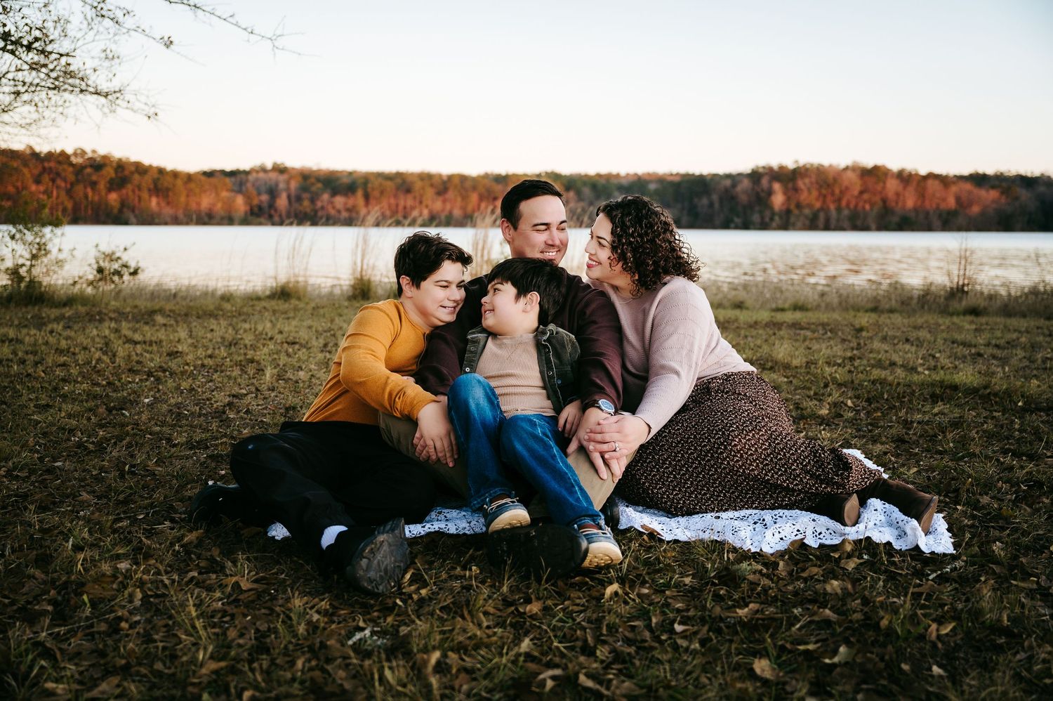 A couple lies together on a blanket in a grassy field during autumn with fall foliage in the background.