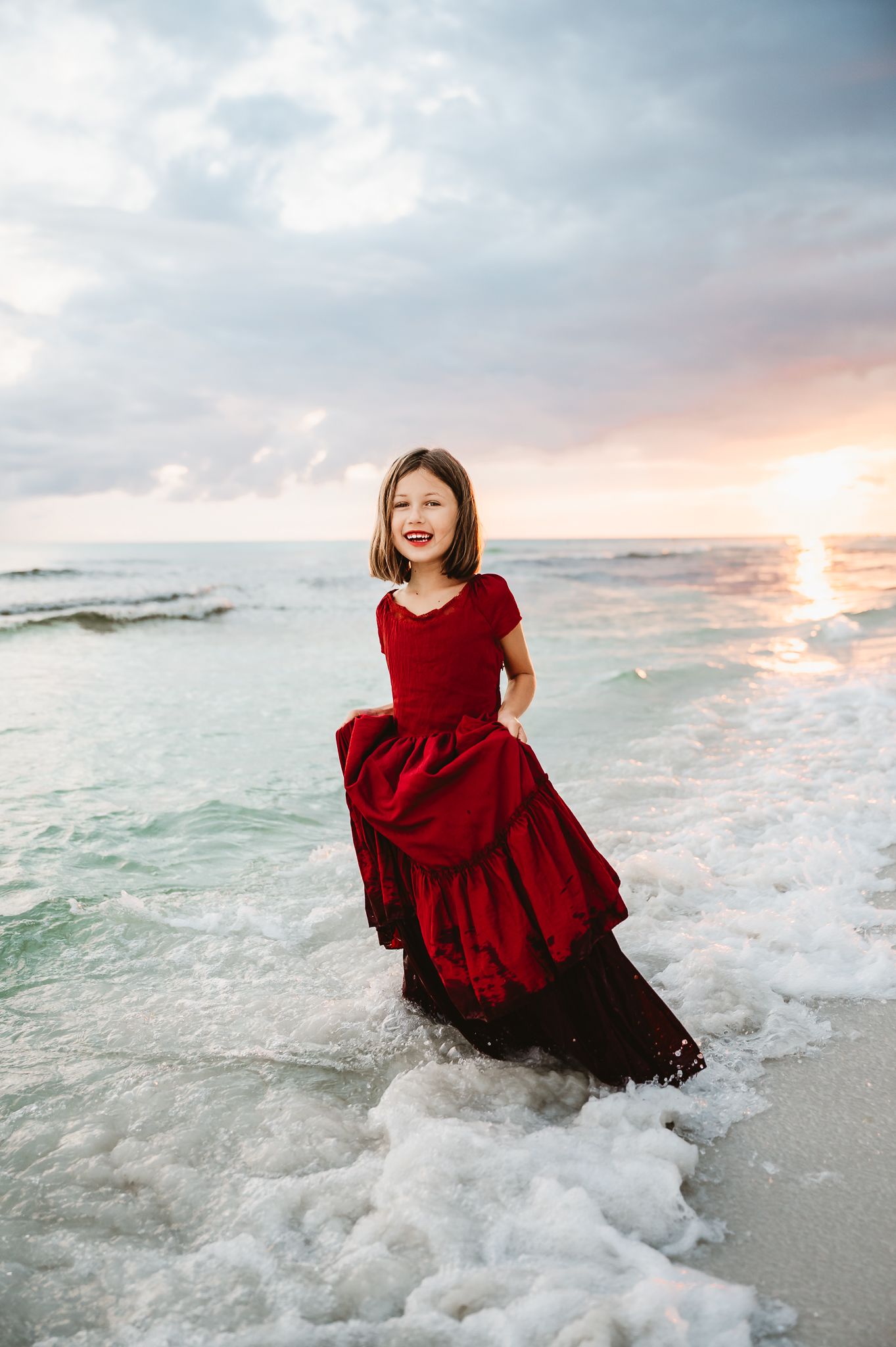 A figure in a flowing red dress stands in the ocean waves at sunset creating a dreamy beachside scene.