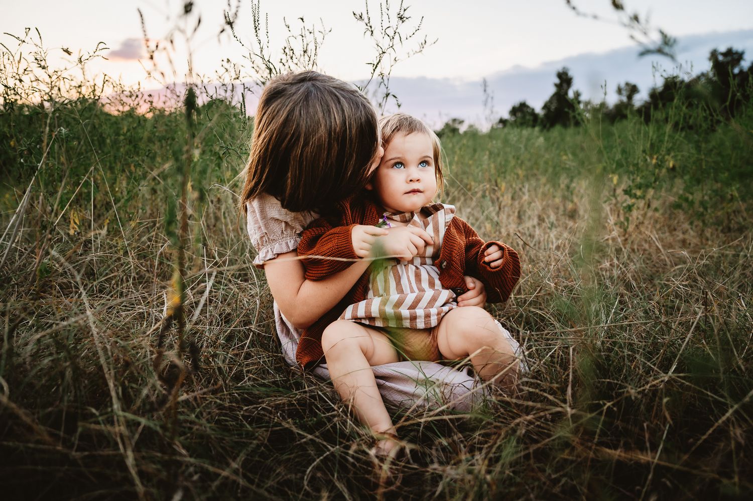 A tender outdoor moment captured in tall grass at sunset with warm earth tones and natural lighting.