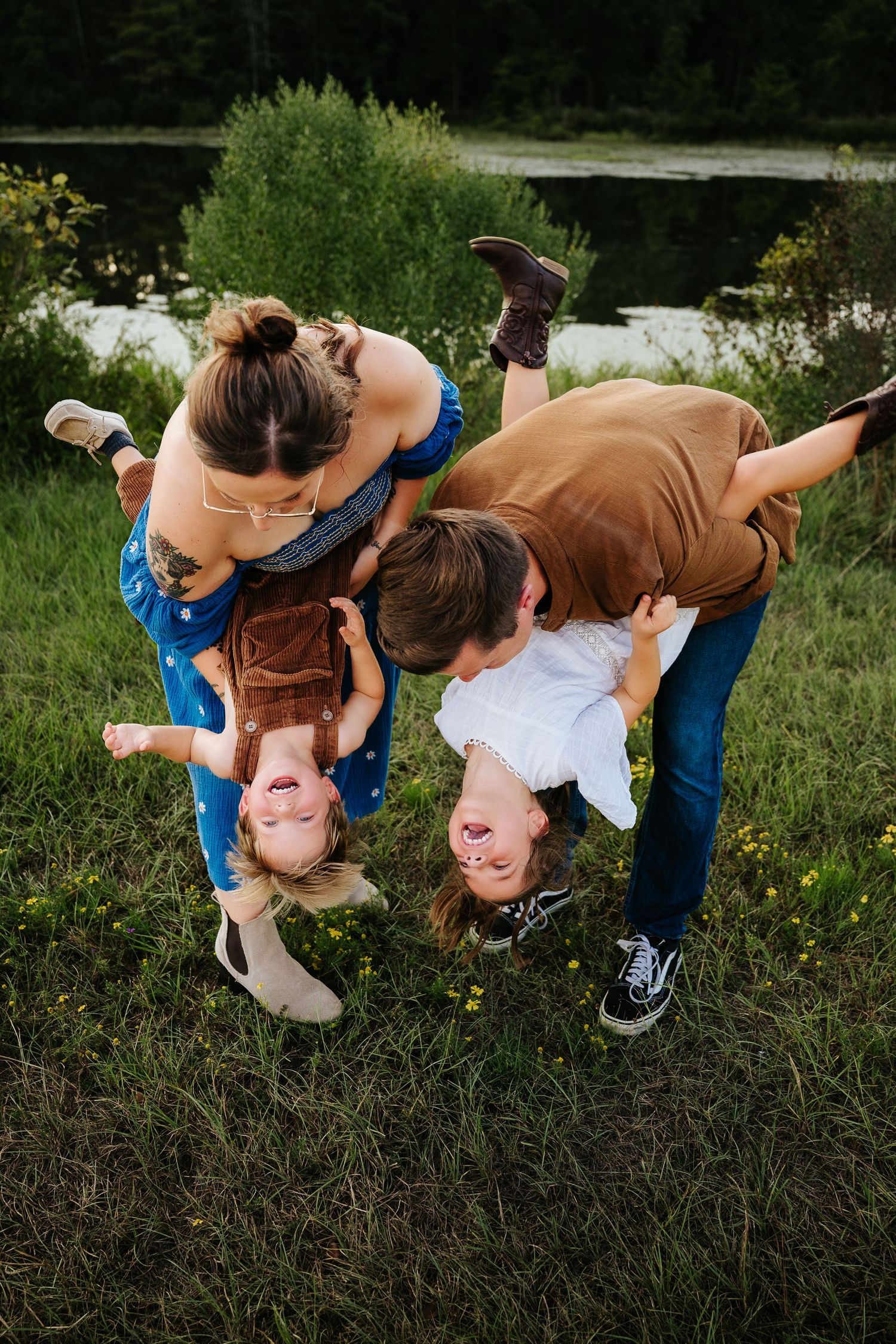 Two parents playfully hold their laughing children upside down outdoors near a pond surrounded by lush green grass.
