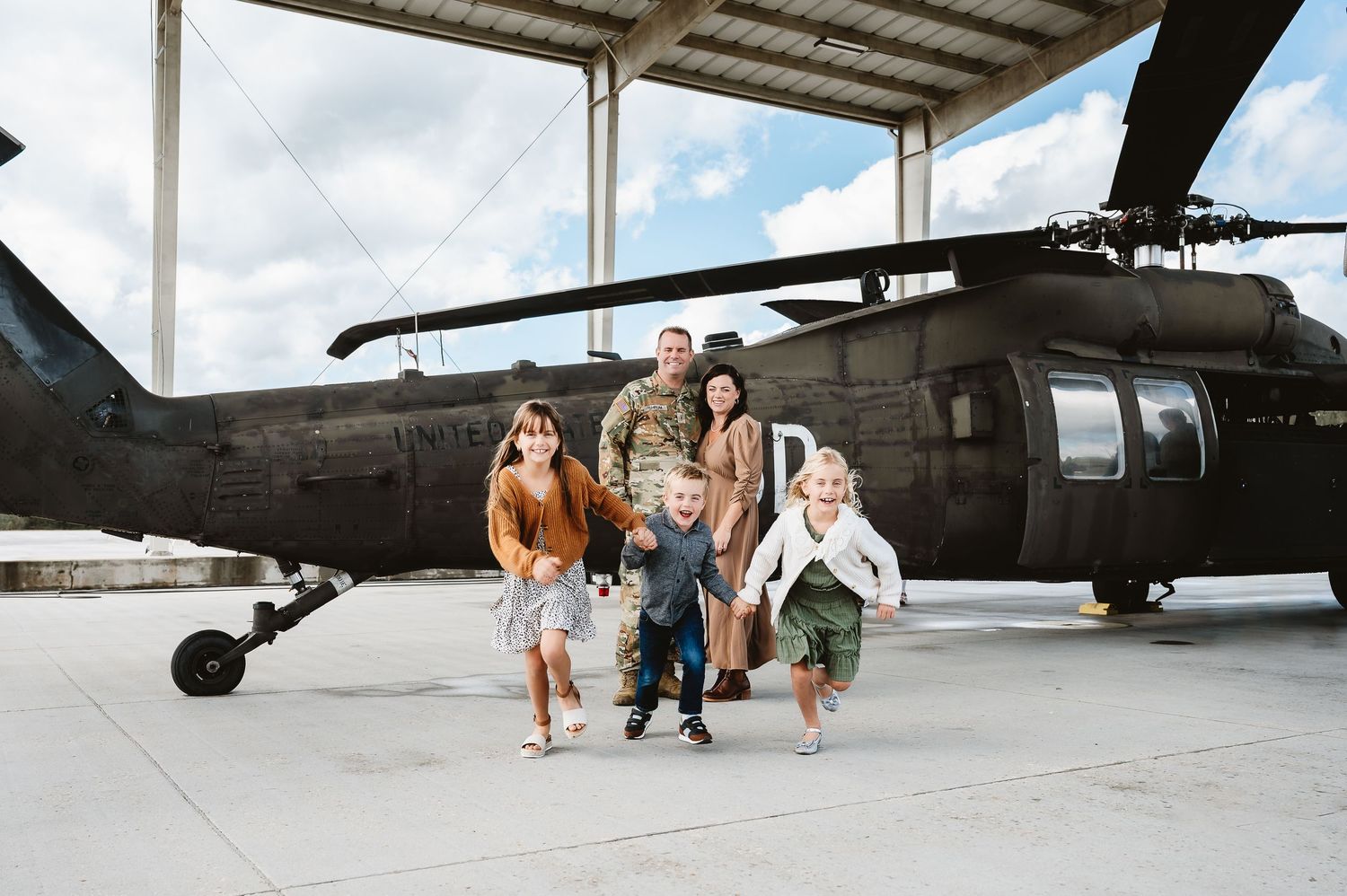 Military helicopter on tarmac with people standing nearby against cloudy sky backdrop.