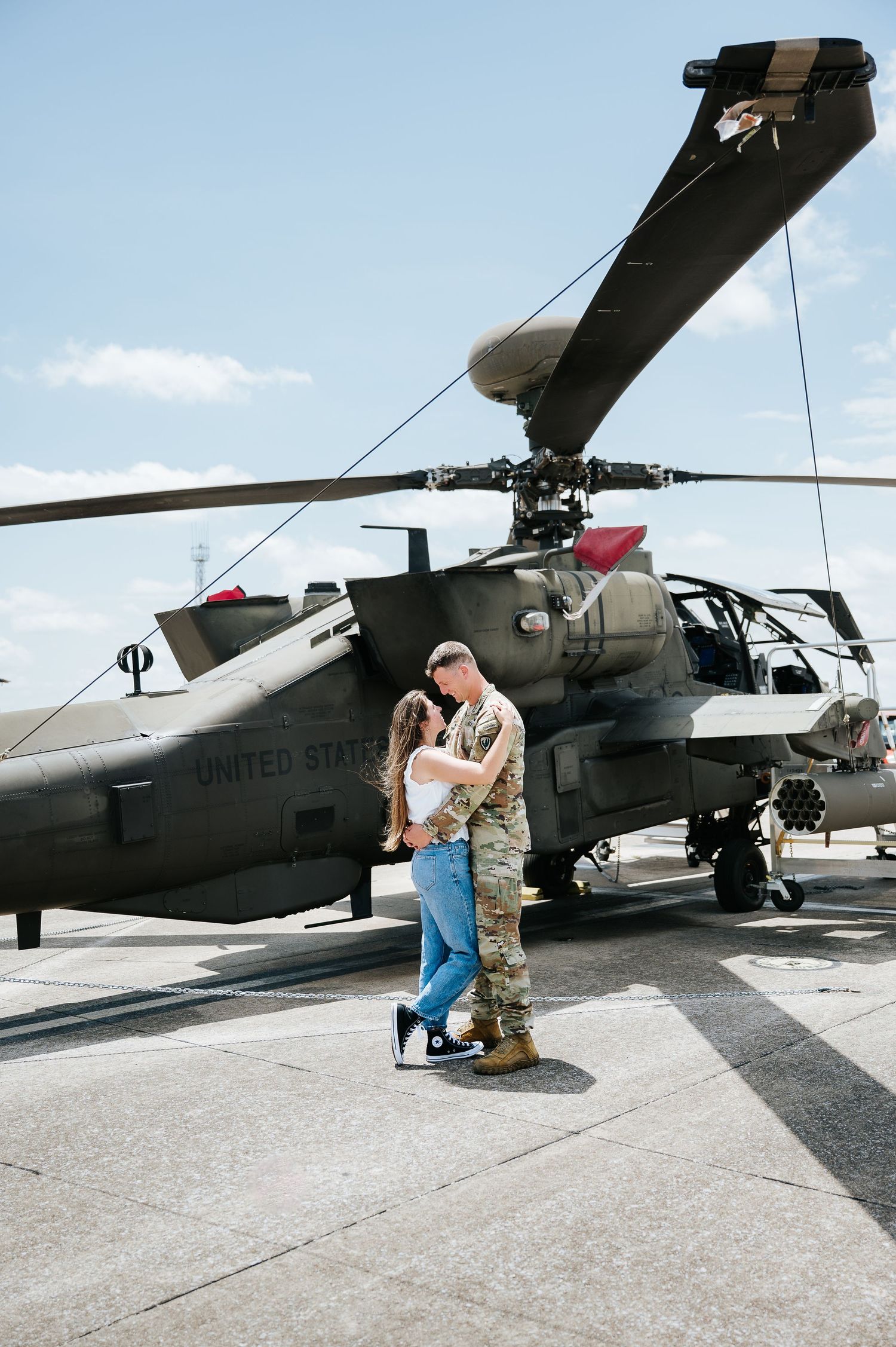 Soldier in camouflage uniform embracing partner in front of a US Army Apache helicopter on a sunny day.