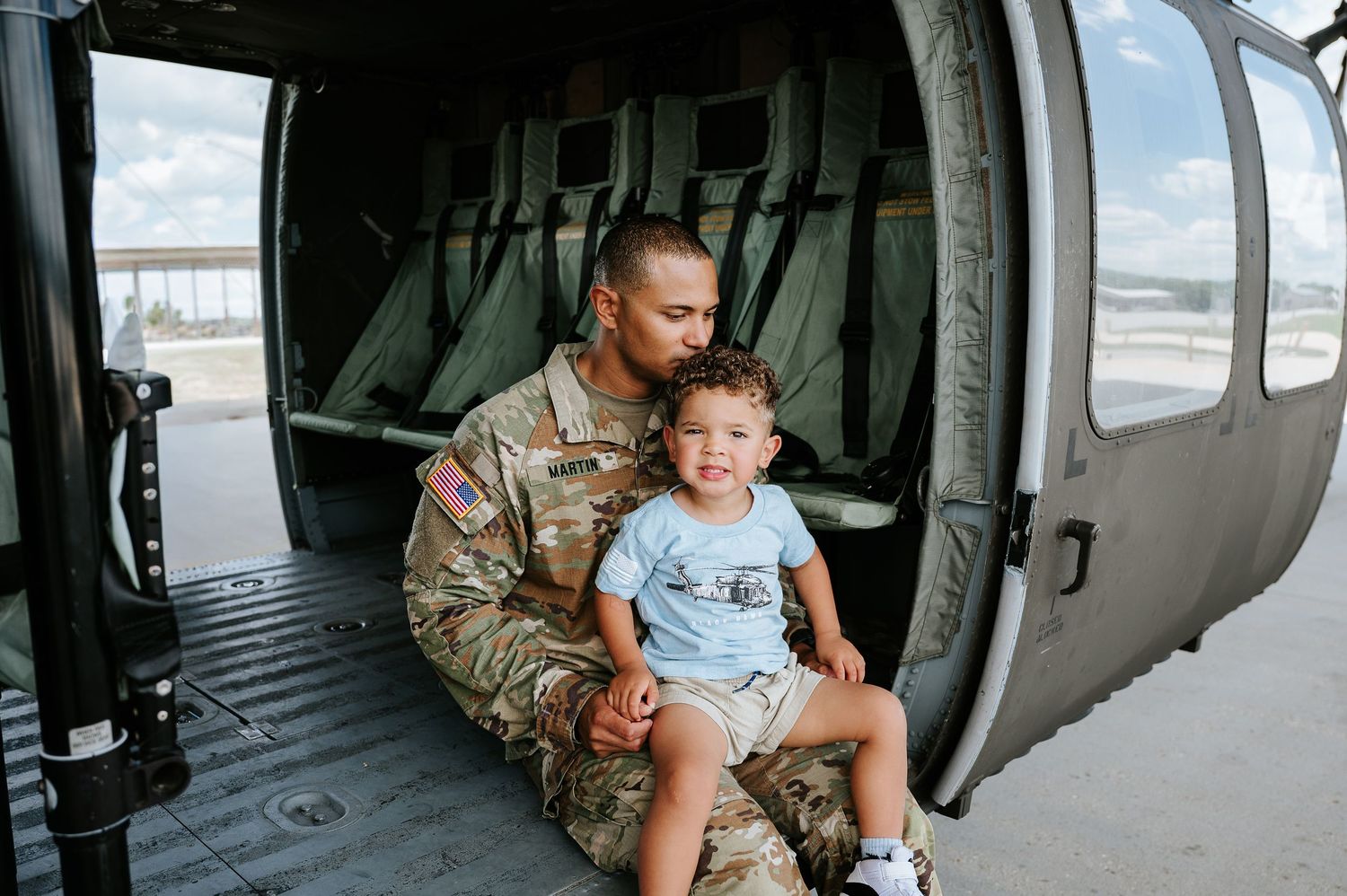 Soldier in camouflage uniform sits with young child inside military helicopter during family visit.
