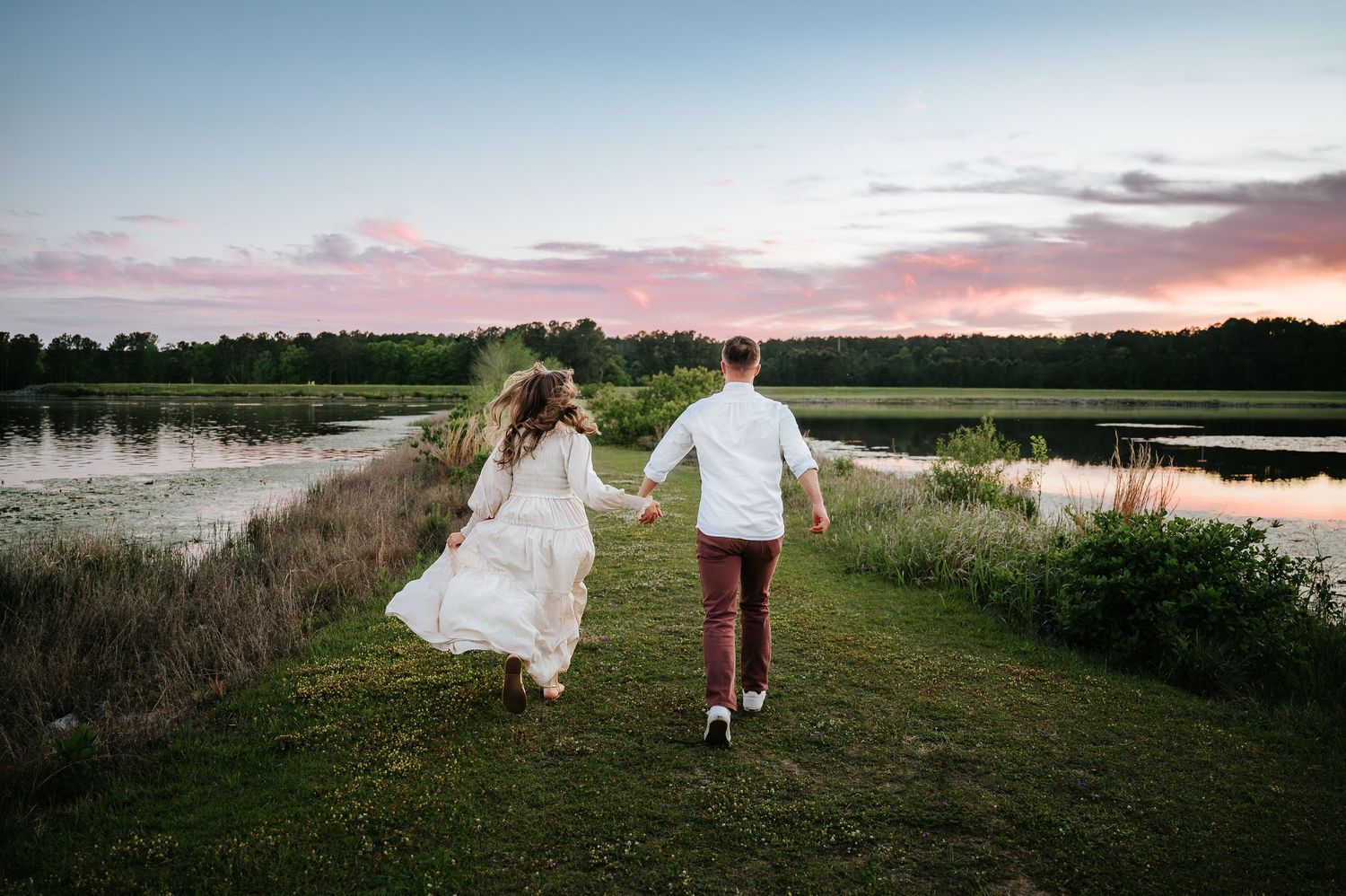 Bride and groom holding hands running by a scenic lake at sunset in a romantic outdoor wedding photo.