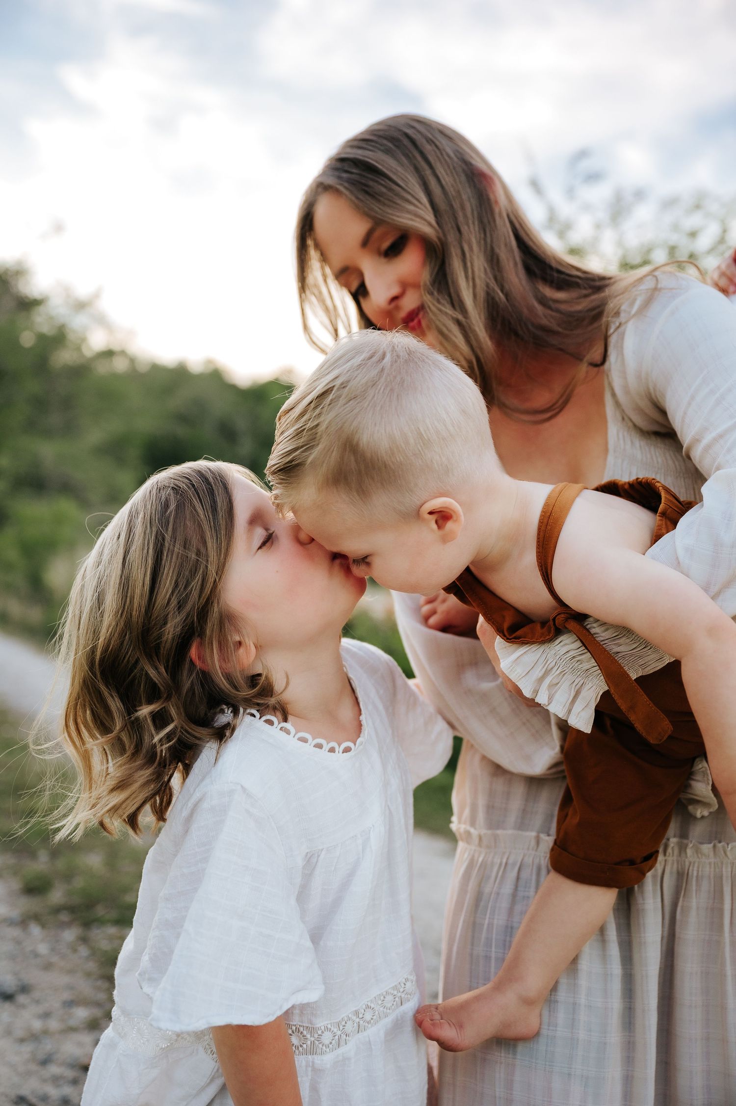 A mother lovingly watches her two young children share a sweet kiss outdoors in soft natural light.