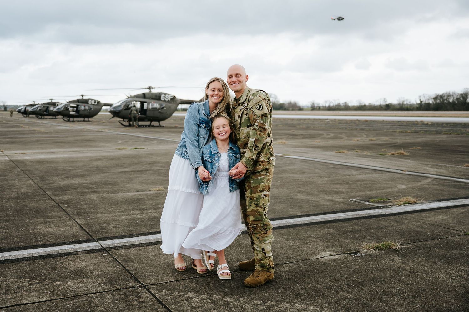 A family stands together on an airfield with military helicopters in the background on an overcast day.