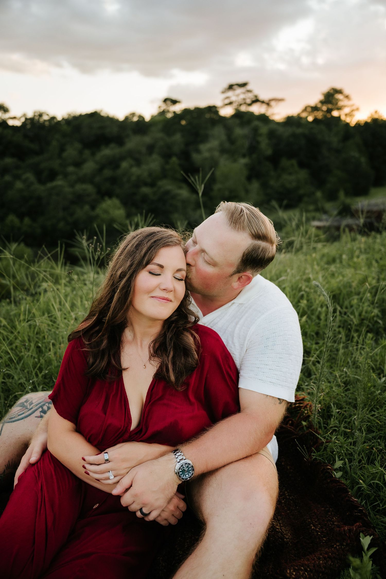 A couple shares a romantic sunset moment outdoors, man kissing woman in red dress, surrounded by lush green fields.