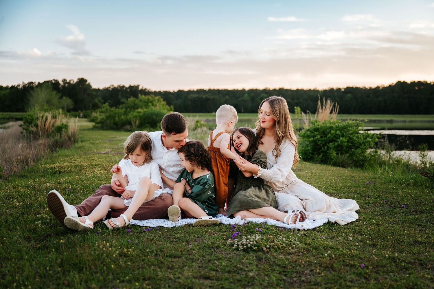 Family of five sitting on a blanket in a lush green field at sunset near a pond.