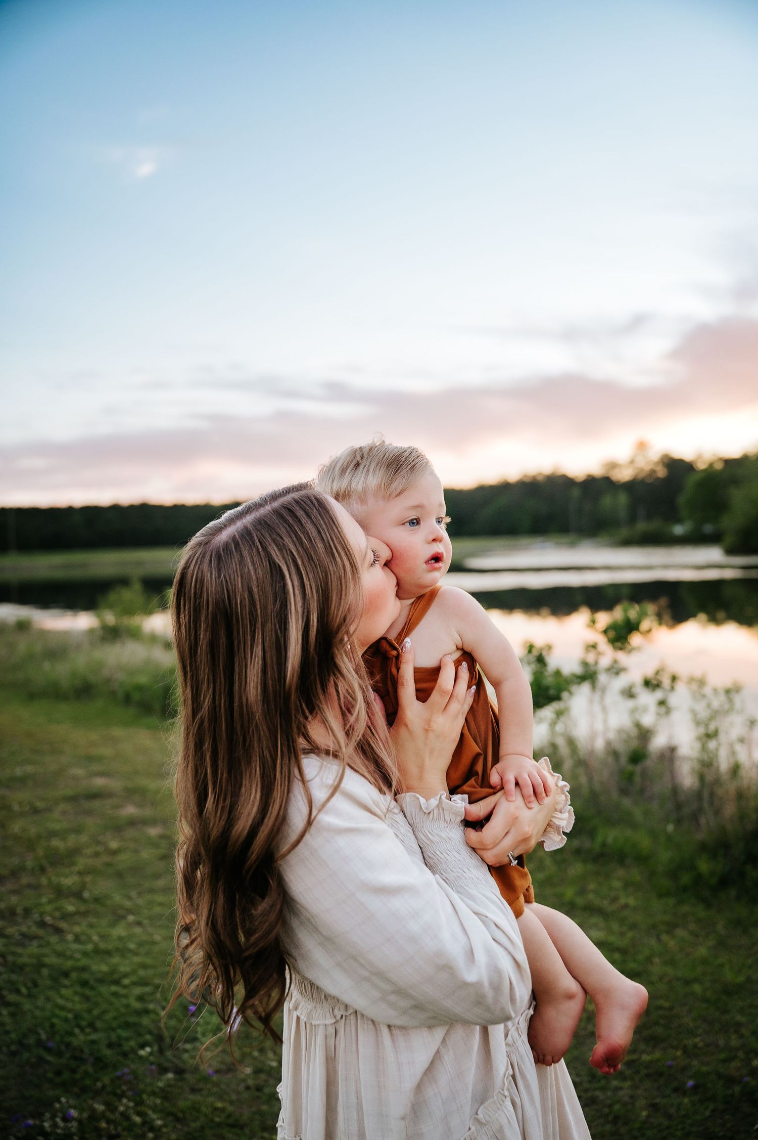 A mother tenderly kisses her toddler while holding them outdoors near a lake at sunset.