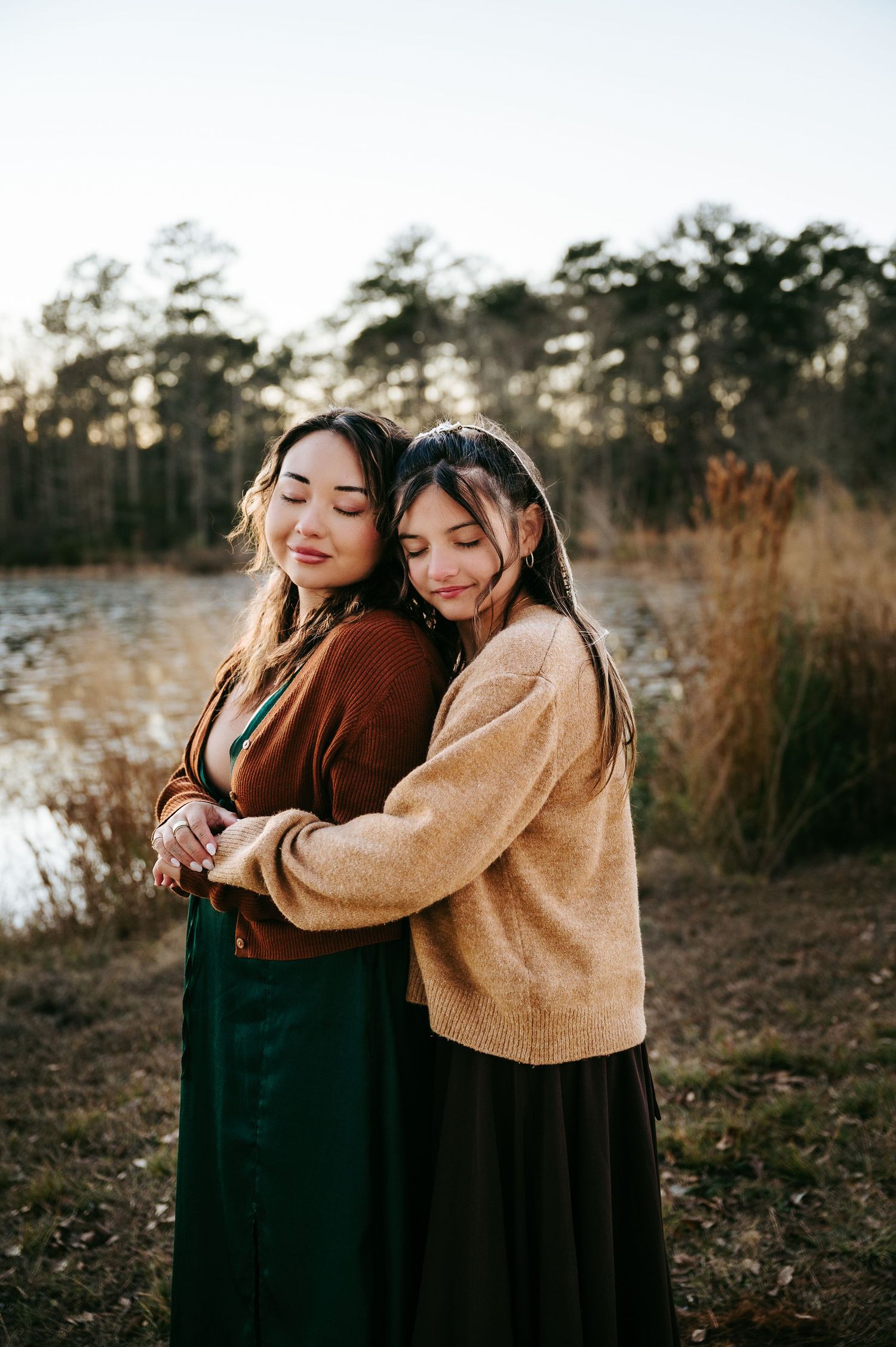 Two people in warm sweaters embrace outdoors during golden hour in a natural setting with tall grass and trees.