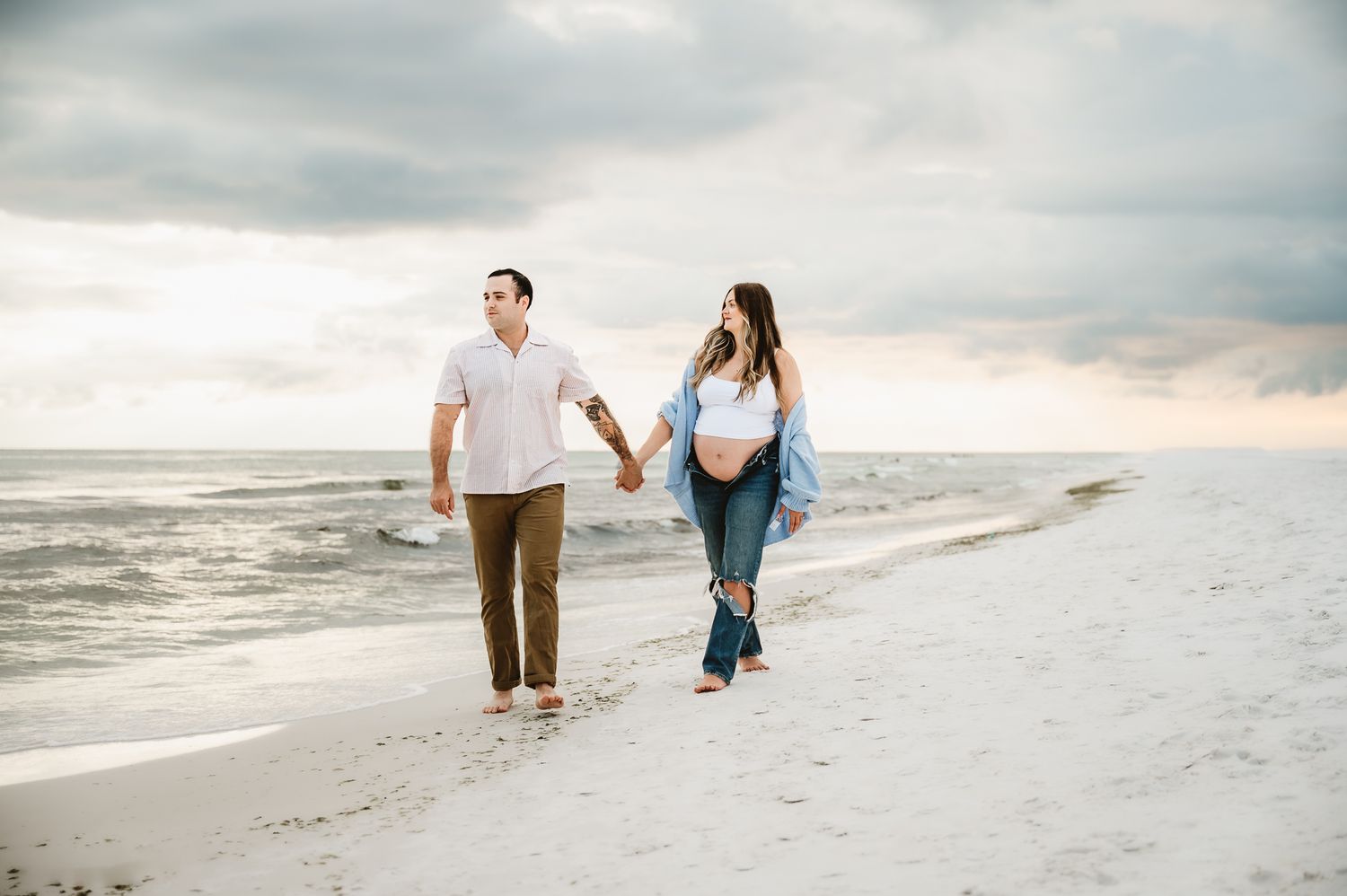 Two people walking hand in hand along a sandy beach at sunset while ocean waves roll in.