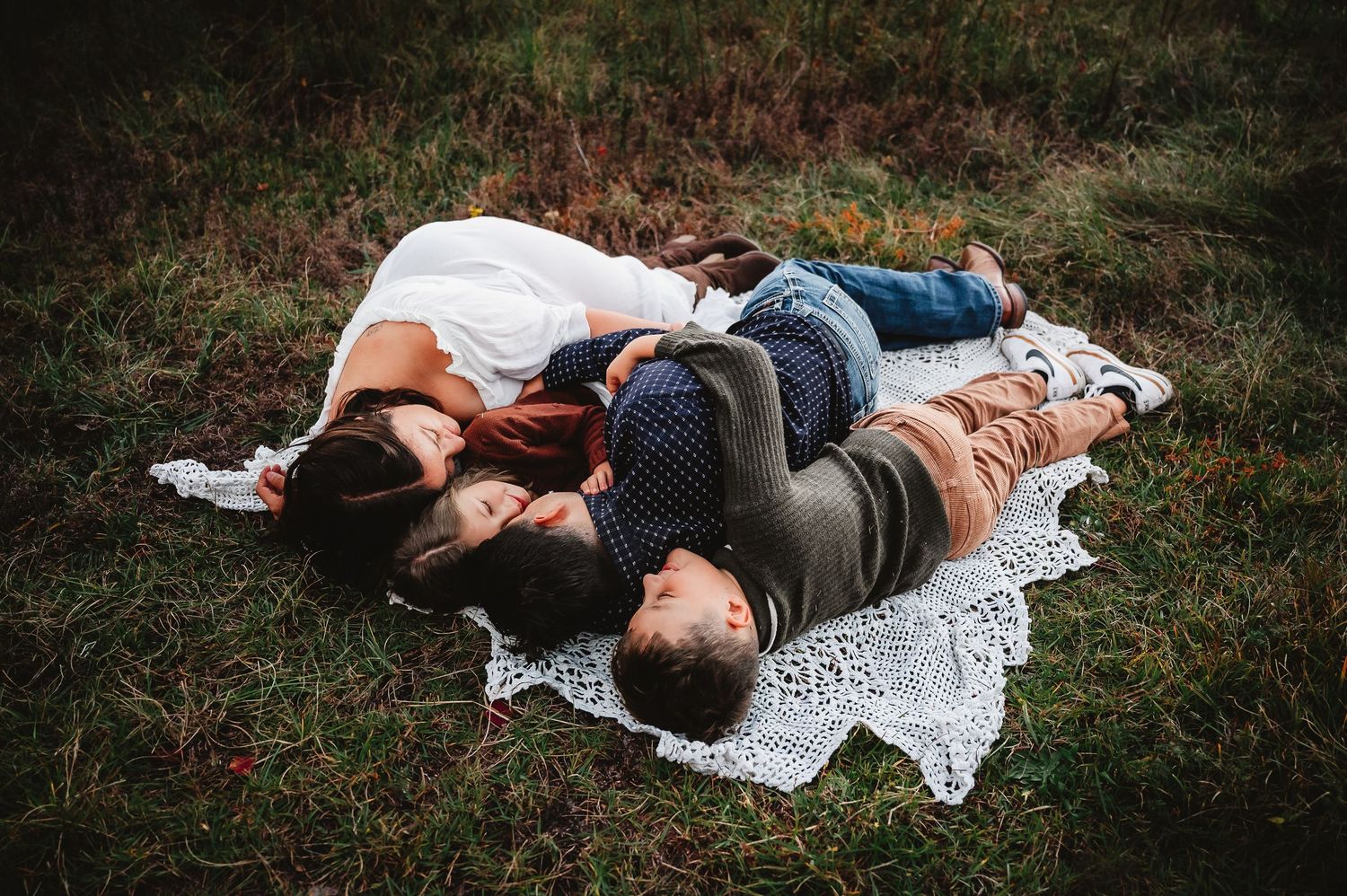 A couple lies together on a white lace blanket in a grassy outdoor setting during a romantic photoshoot.