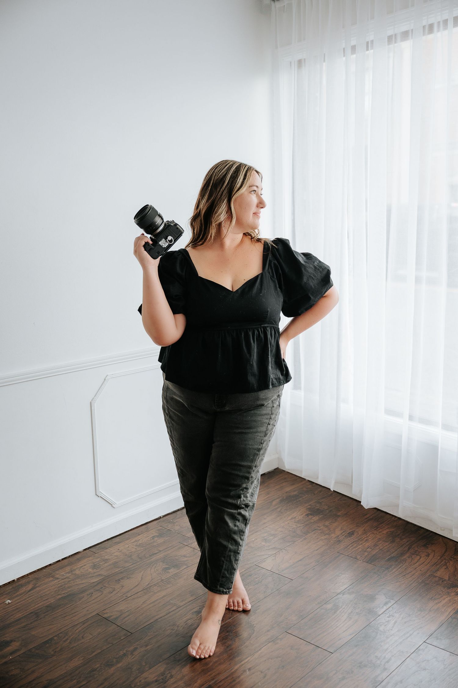 Woman in black puff-sleeve top and olive pants holds camera, posing barefoot in bright white studio room.