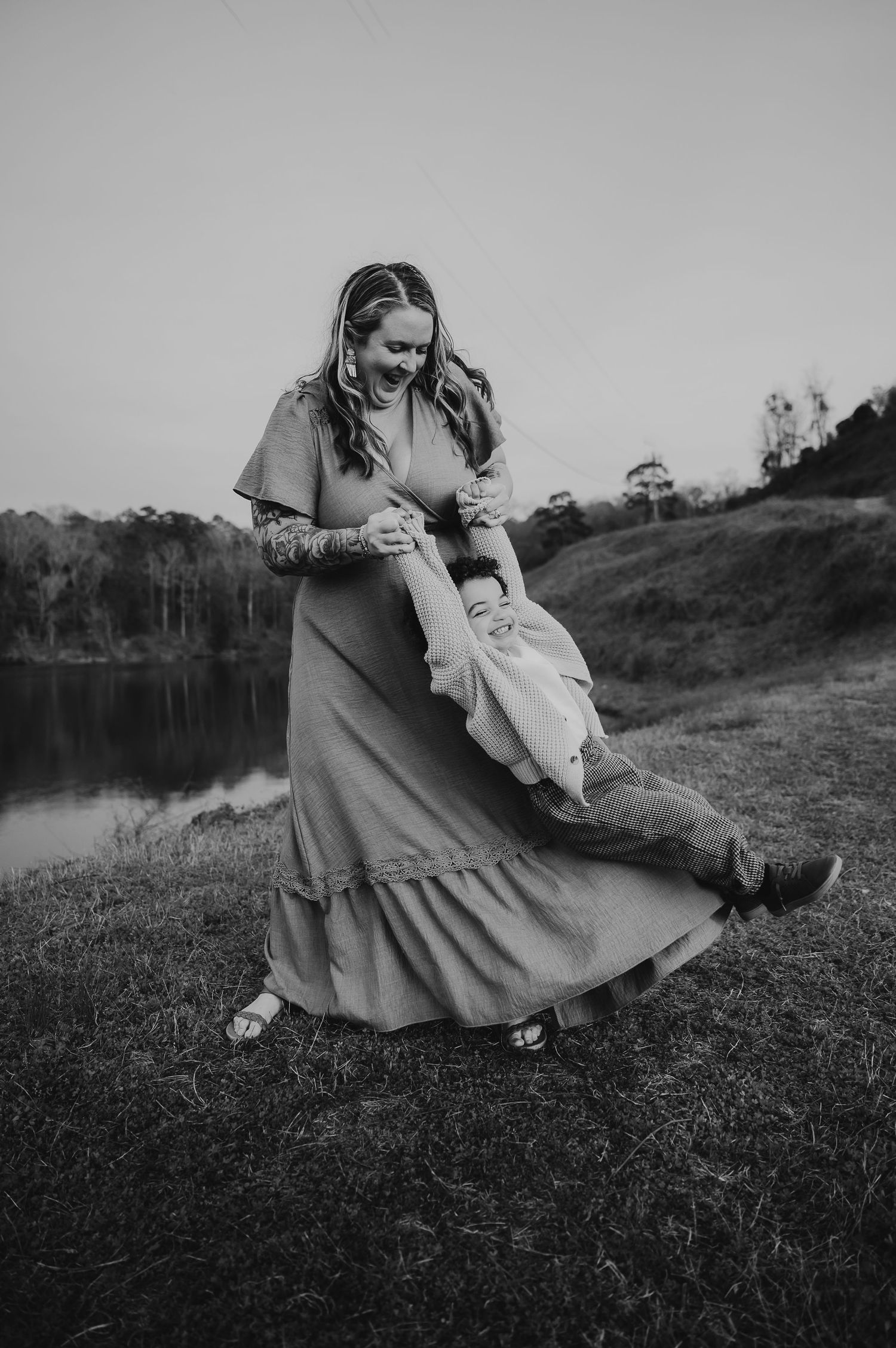 Black and white photo of a laughing woman in a maxi dress swinging a child outdoors near a lake.