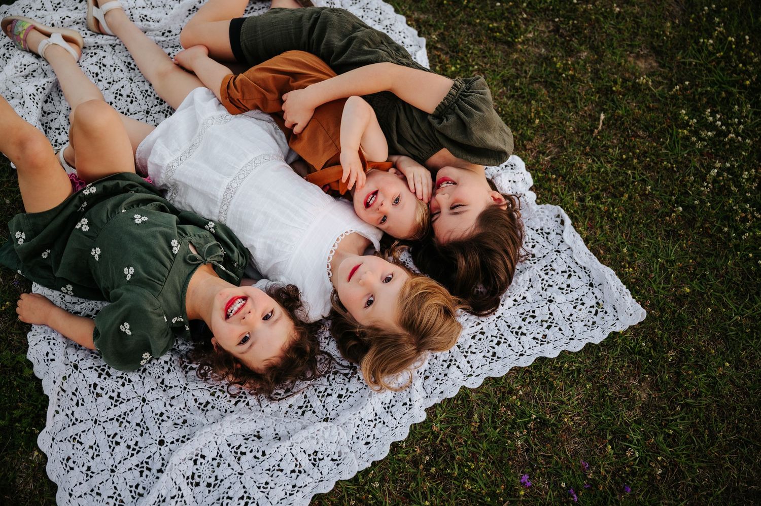 Four women lying on a white lace blanket on green grass, smiling and posing together for a family portrait.