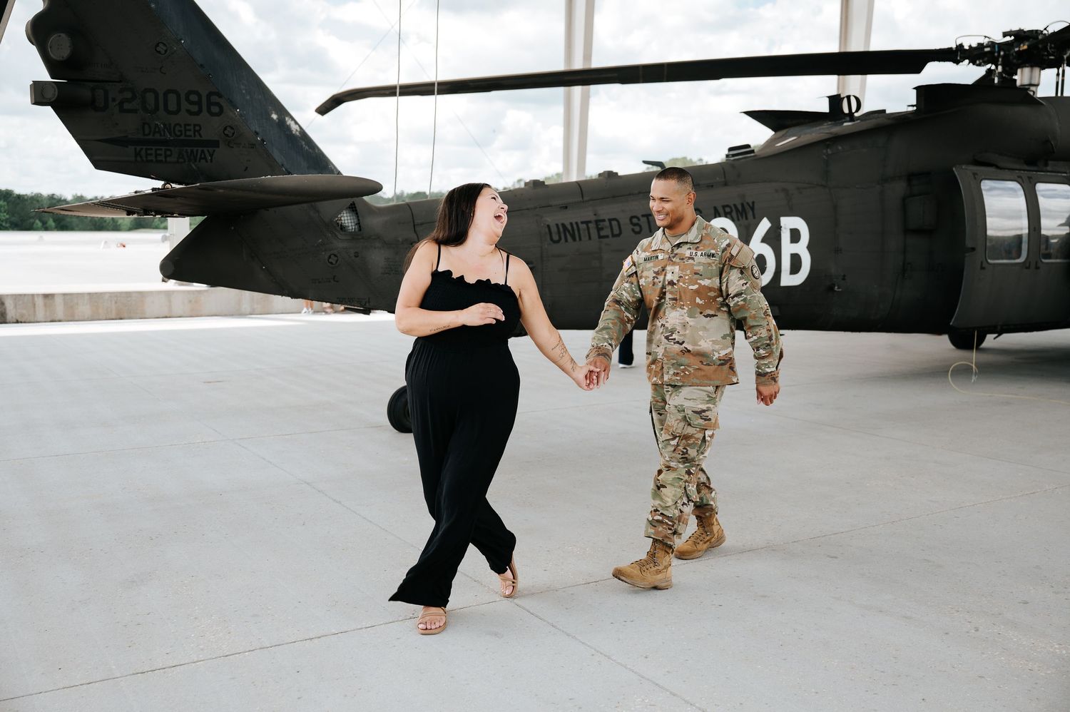 Military couple holding hands and walking in front of a UH-60 Black Hawk helicopter on a tarmac.