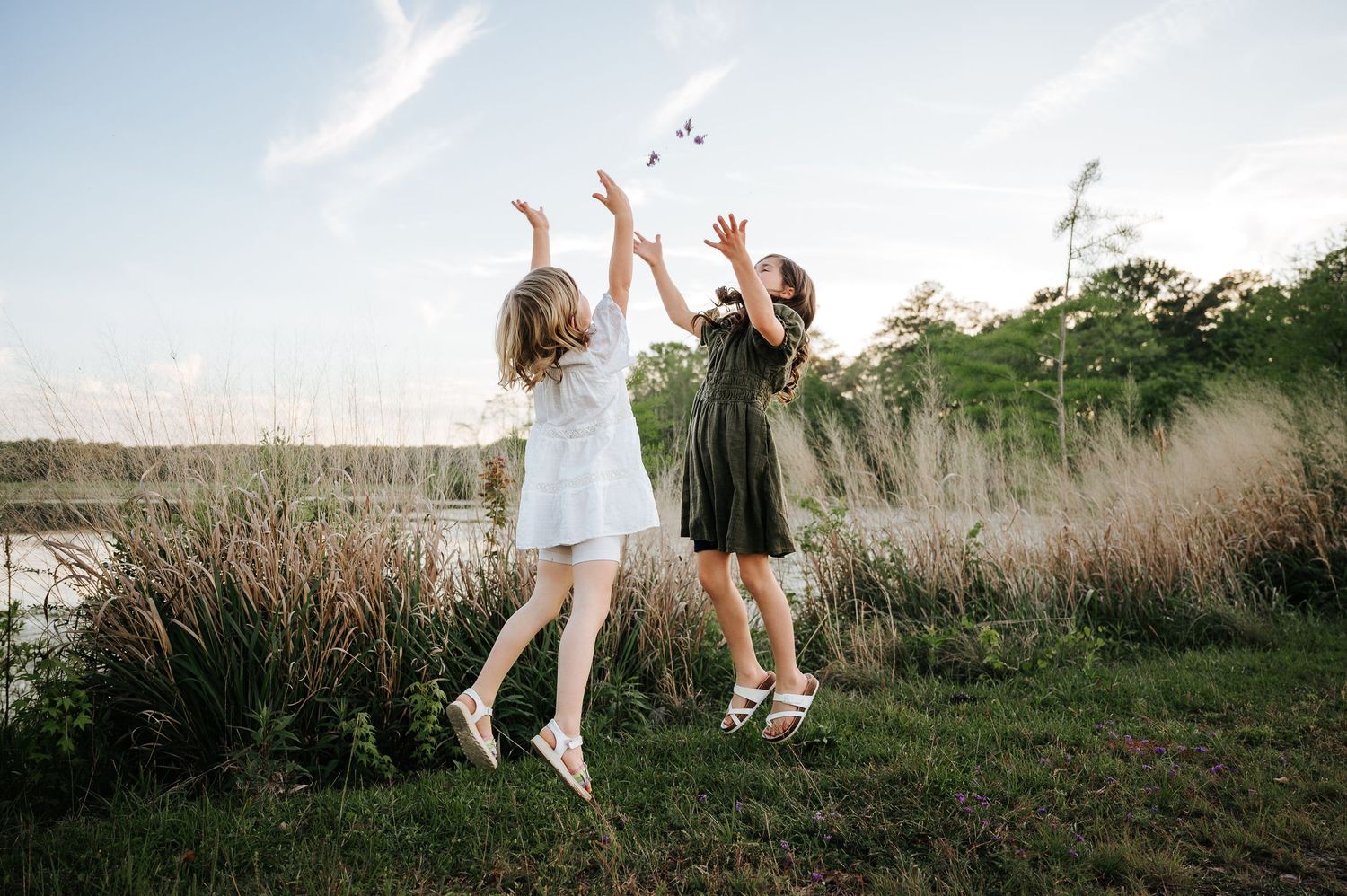 Two young girls in dresses joyfully jump and play outdoors near a serene lake surrounded by tall grass.