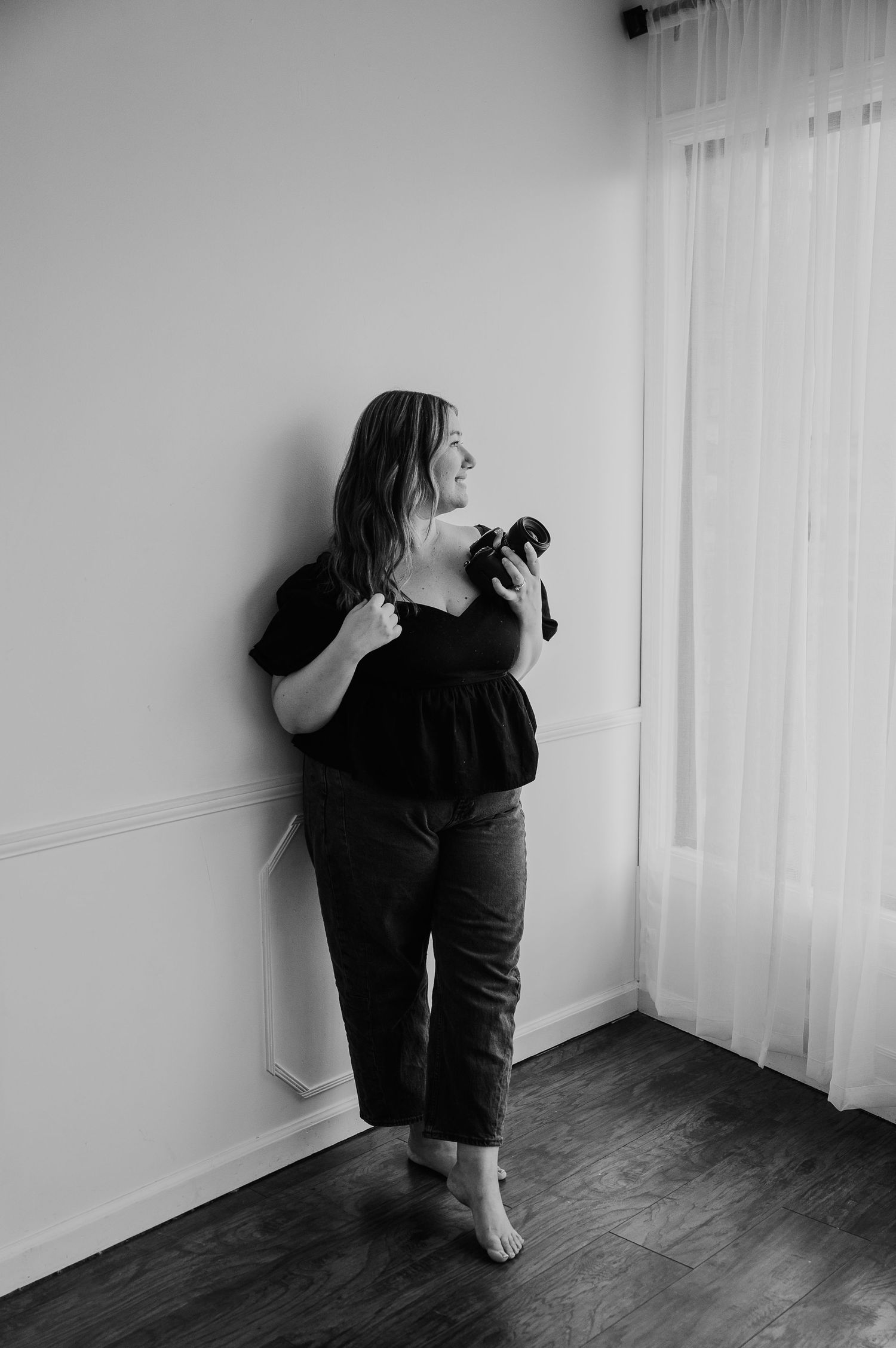 Black and white photo of a woman leaning against a wall holding a camera, wearing a black top and dark pants.