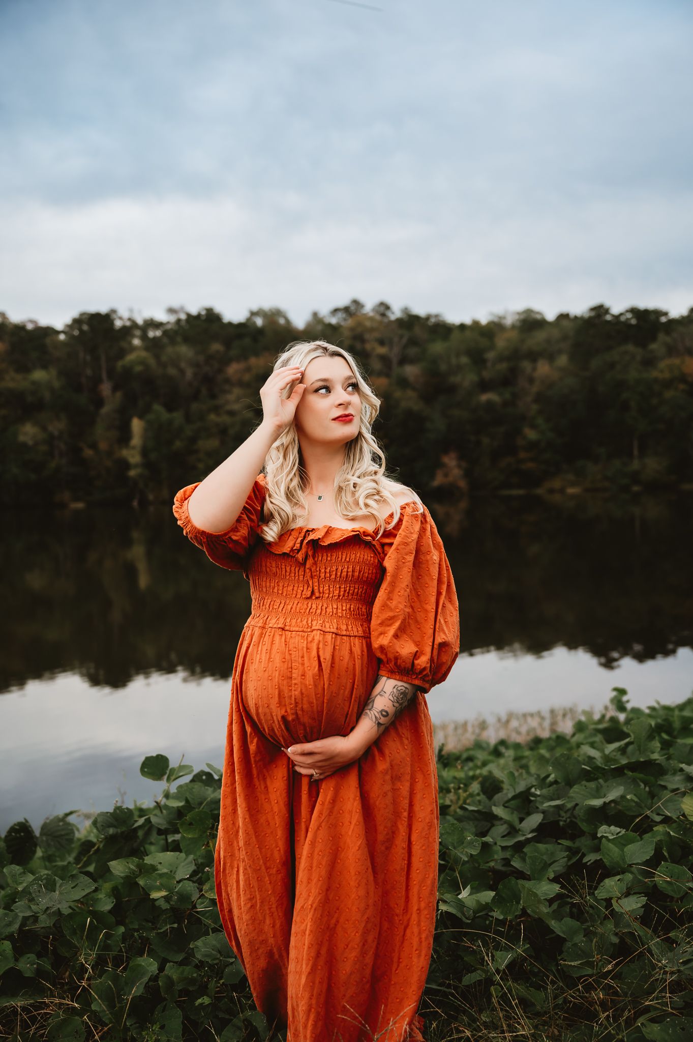 Expectant woman in flowing rust-colored dress stands by tranquil lake at sunset with forest backdrop.