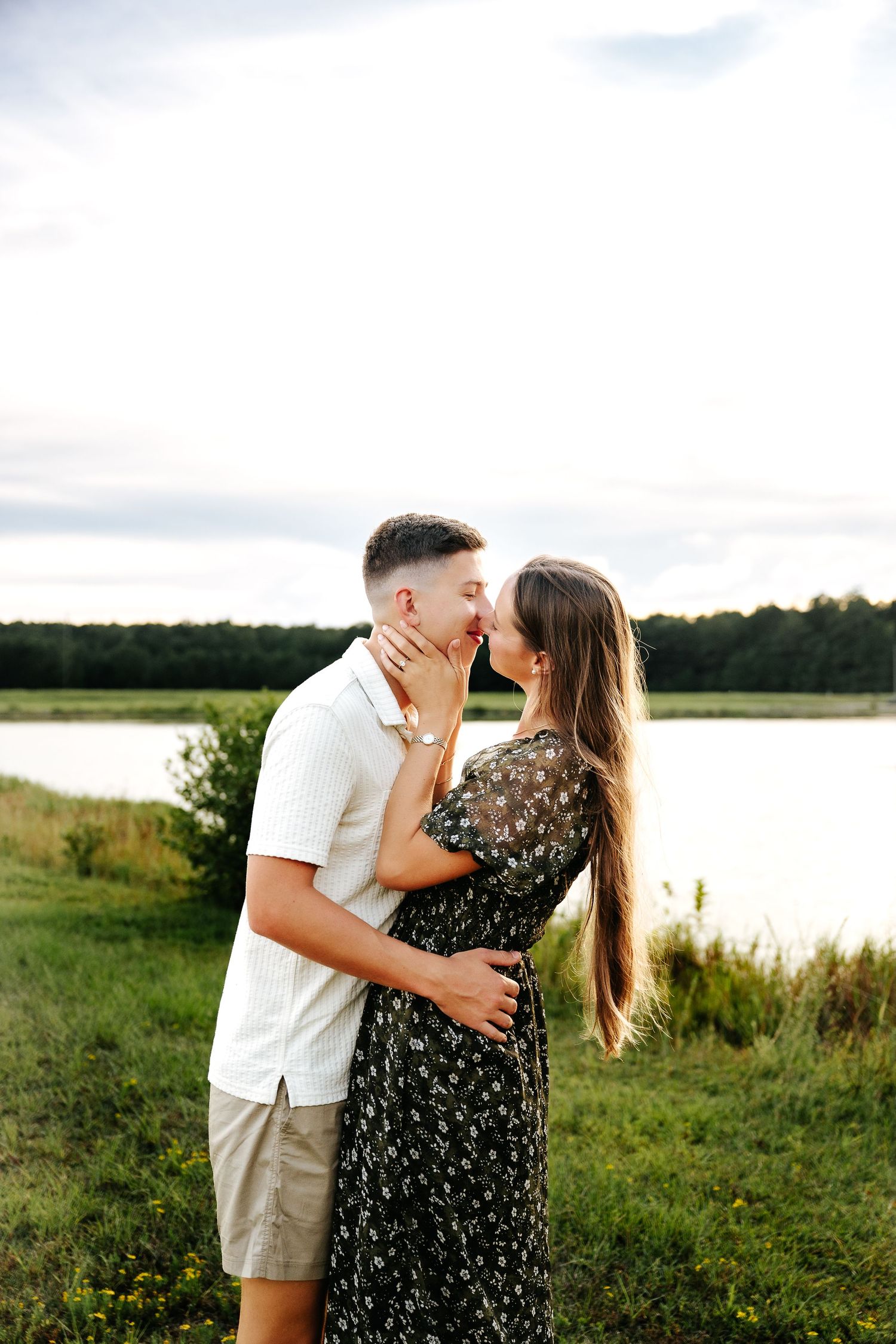 Couple sharing a romantic moment near a serene lake, surrounded by lush greenery at golden hour.