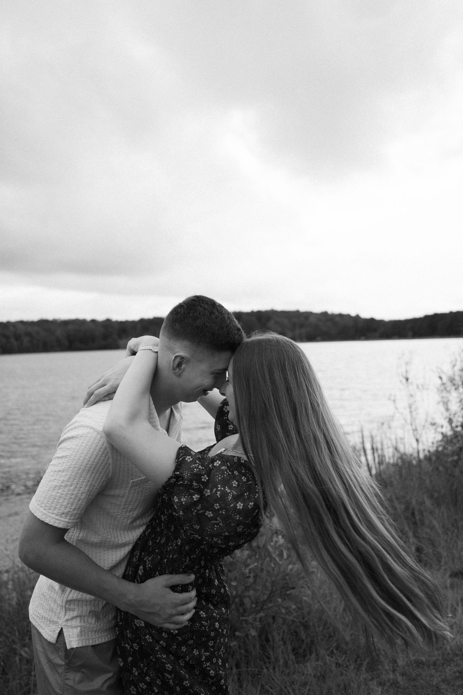Black and white photo of a romantic couple embracing and kissing near a lake with cloudy skies.