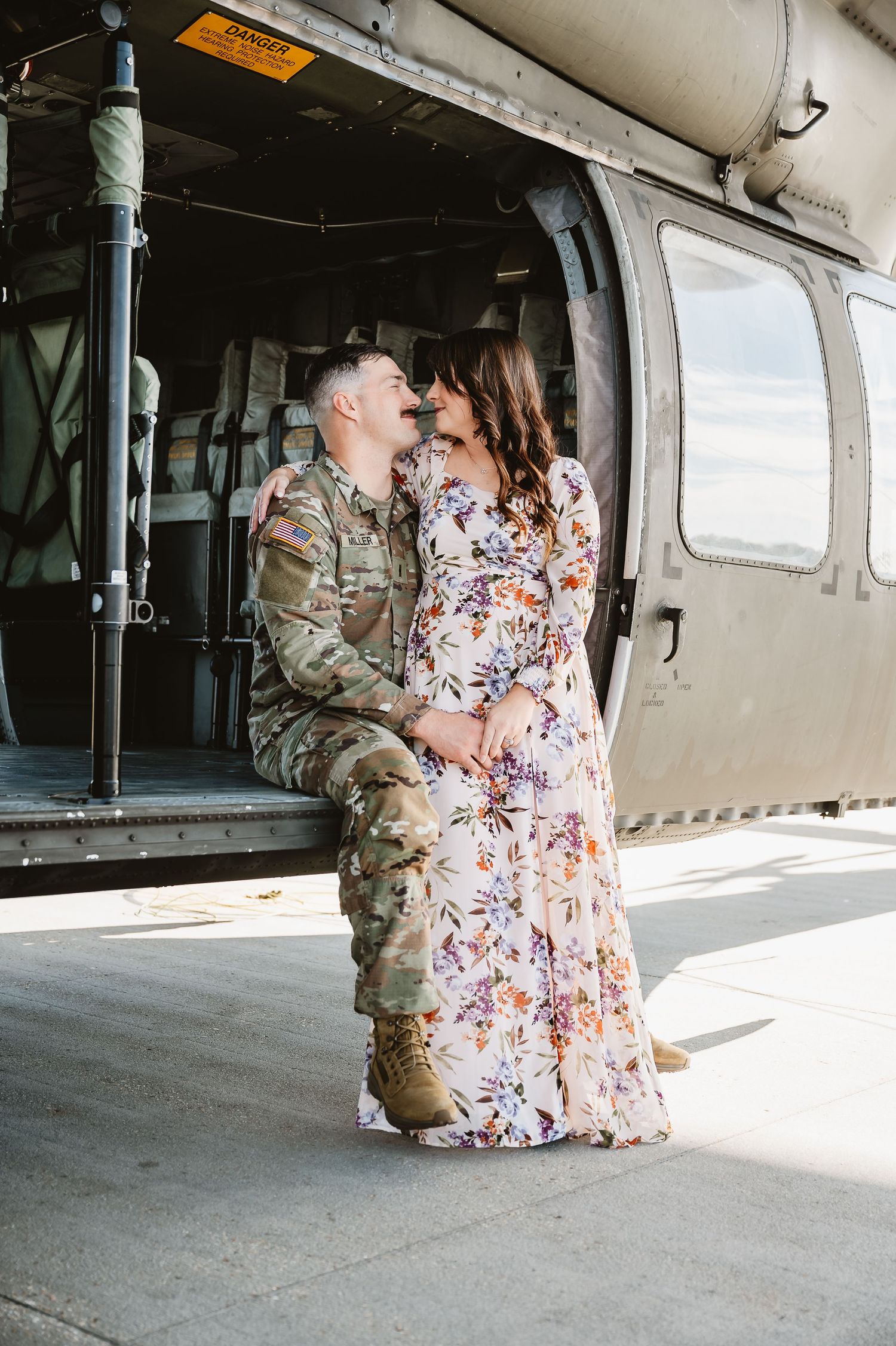 A couple shares an intimate moment in front of a military helicopter, with one person wearing camouflage fatigues.
