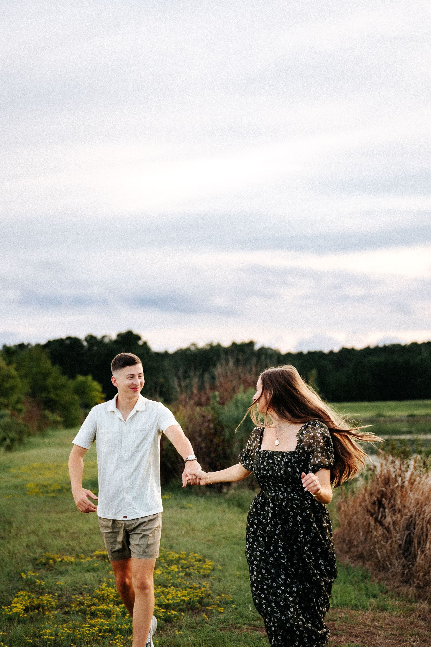 A couple holds hands walking through a green field at dusk, woman in floral dress, man in white shirt and shorts.