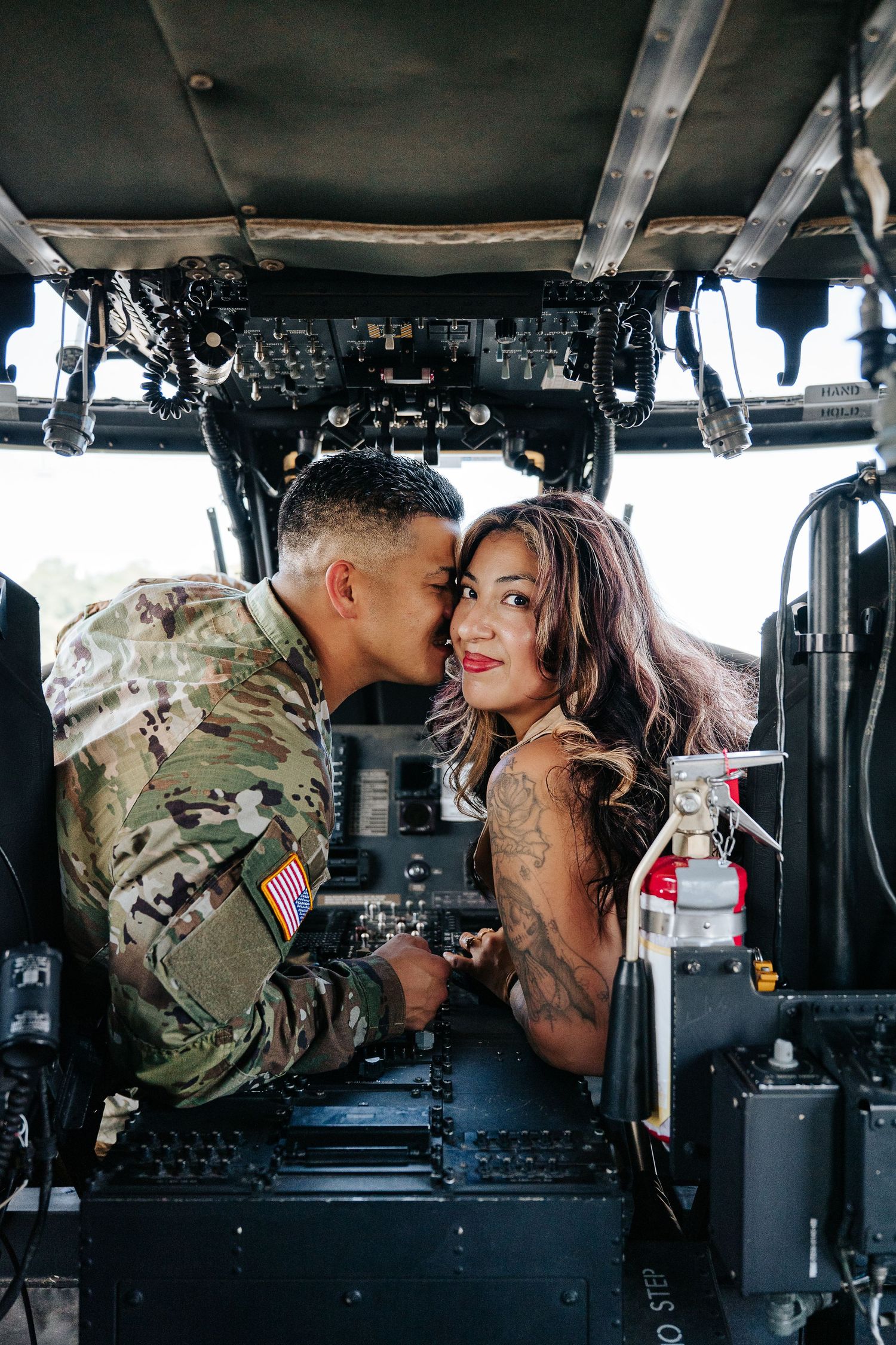 A soldier in camouflage uniform kisses a tattooed woman inside a military helicopter cockpit.