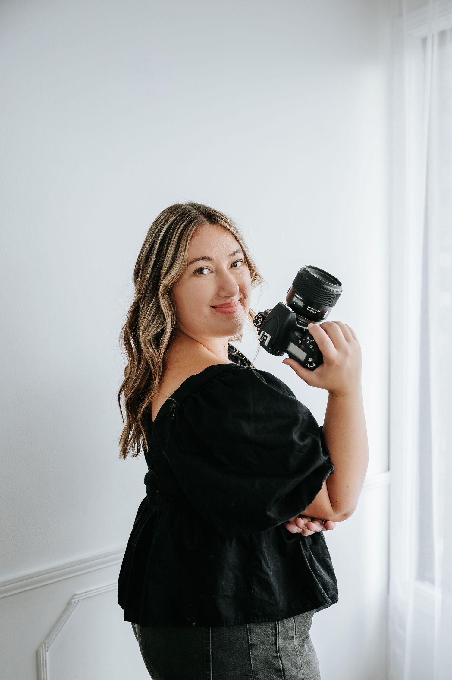 Woman photographer smiling in black off-shoulder top holding DSLR camera in bright white studio.