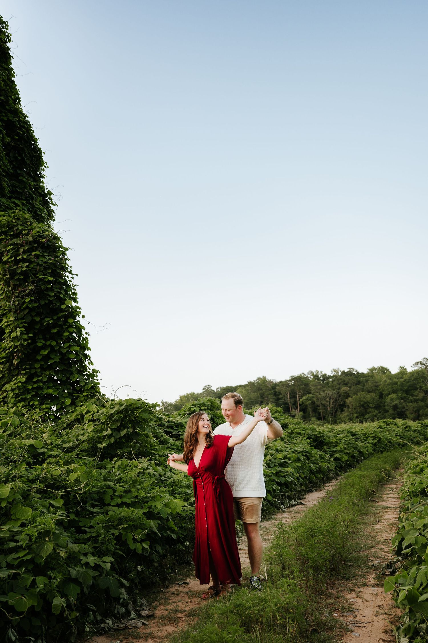 Couple dancing together in a lush green farm field, woman in red dress, man in white shirt, clear blue sky.