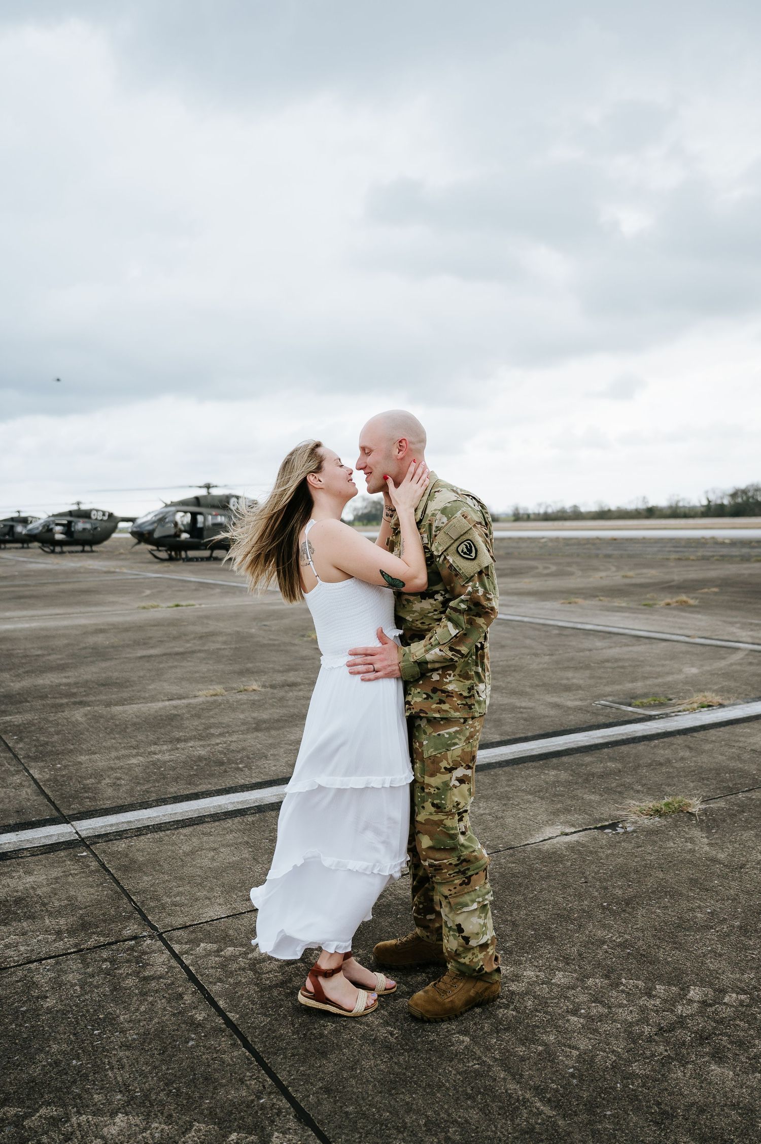 A tender moment shared between a service member in camouflage uniform and someone in a white dress on an empty airfield.
