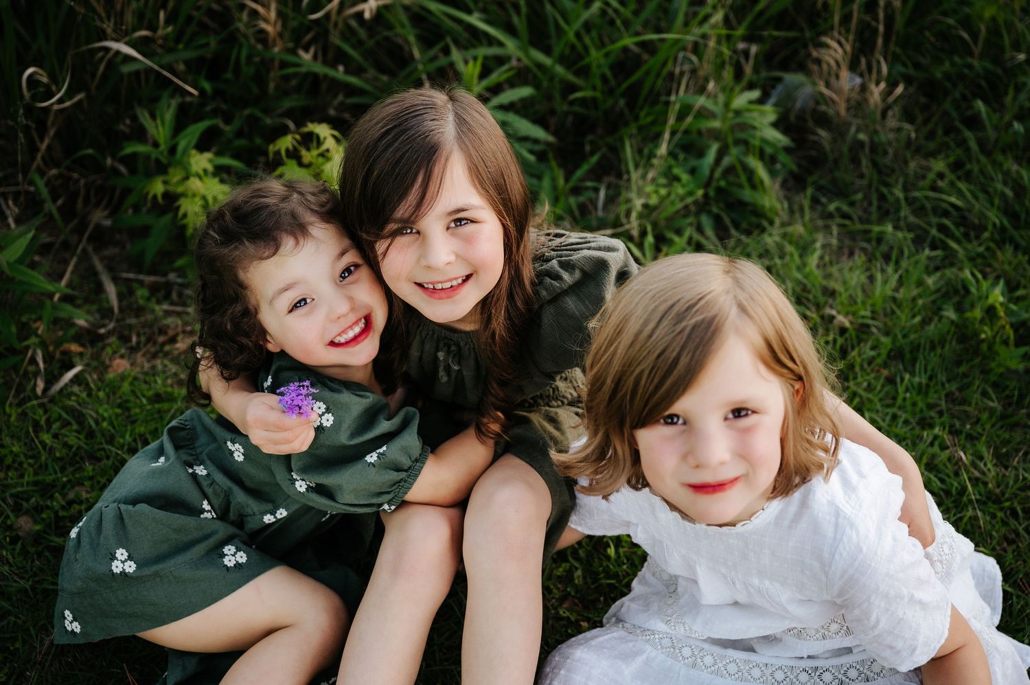 Three smiling young girls sitting together outdoors in green dresses, hugging and posing for a portrait photo.