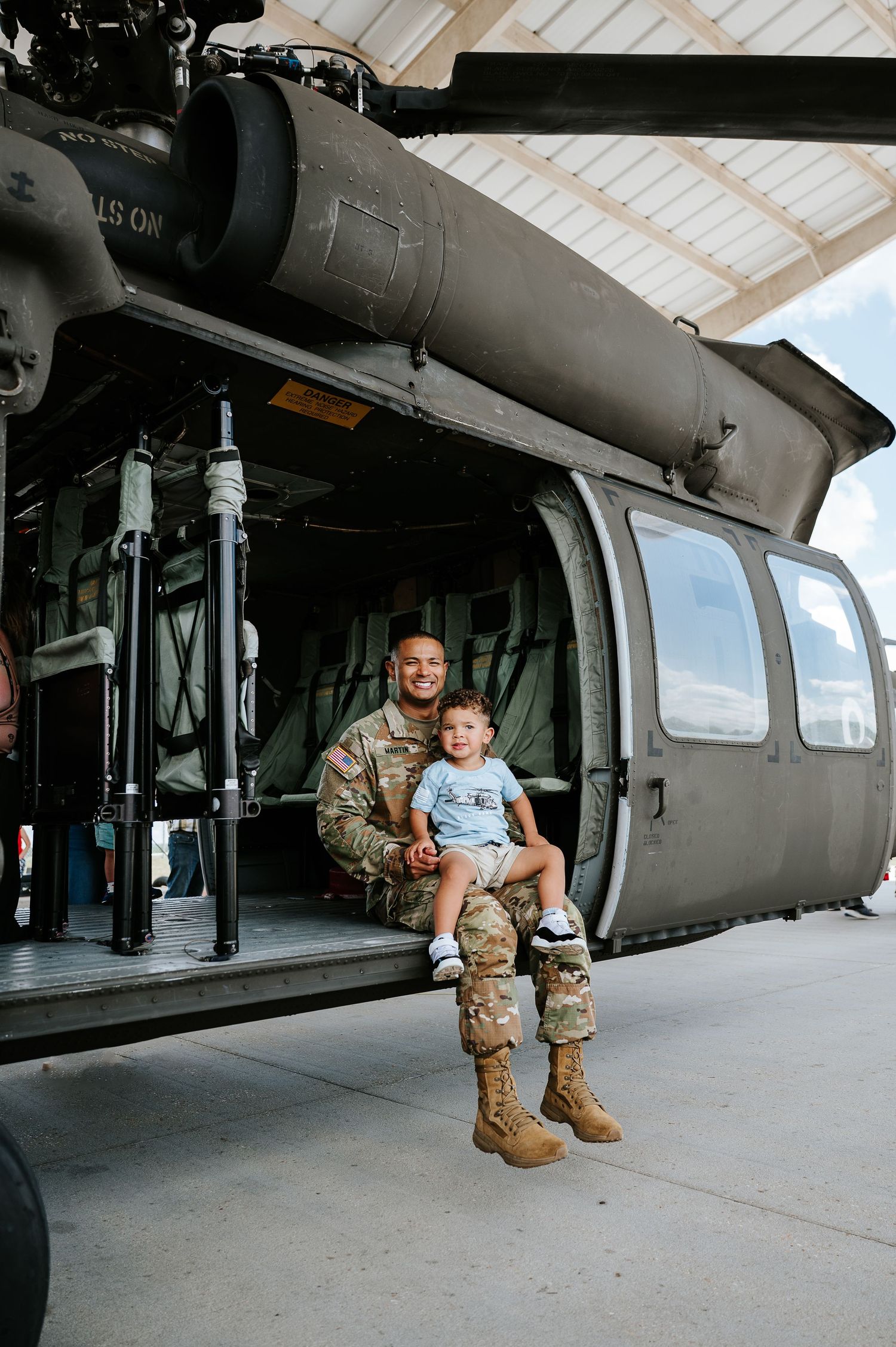 Military soldier in uniform sits with young child in front of open helicopter in hangar.