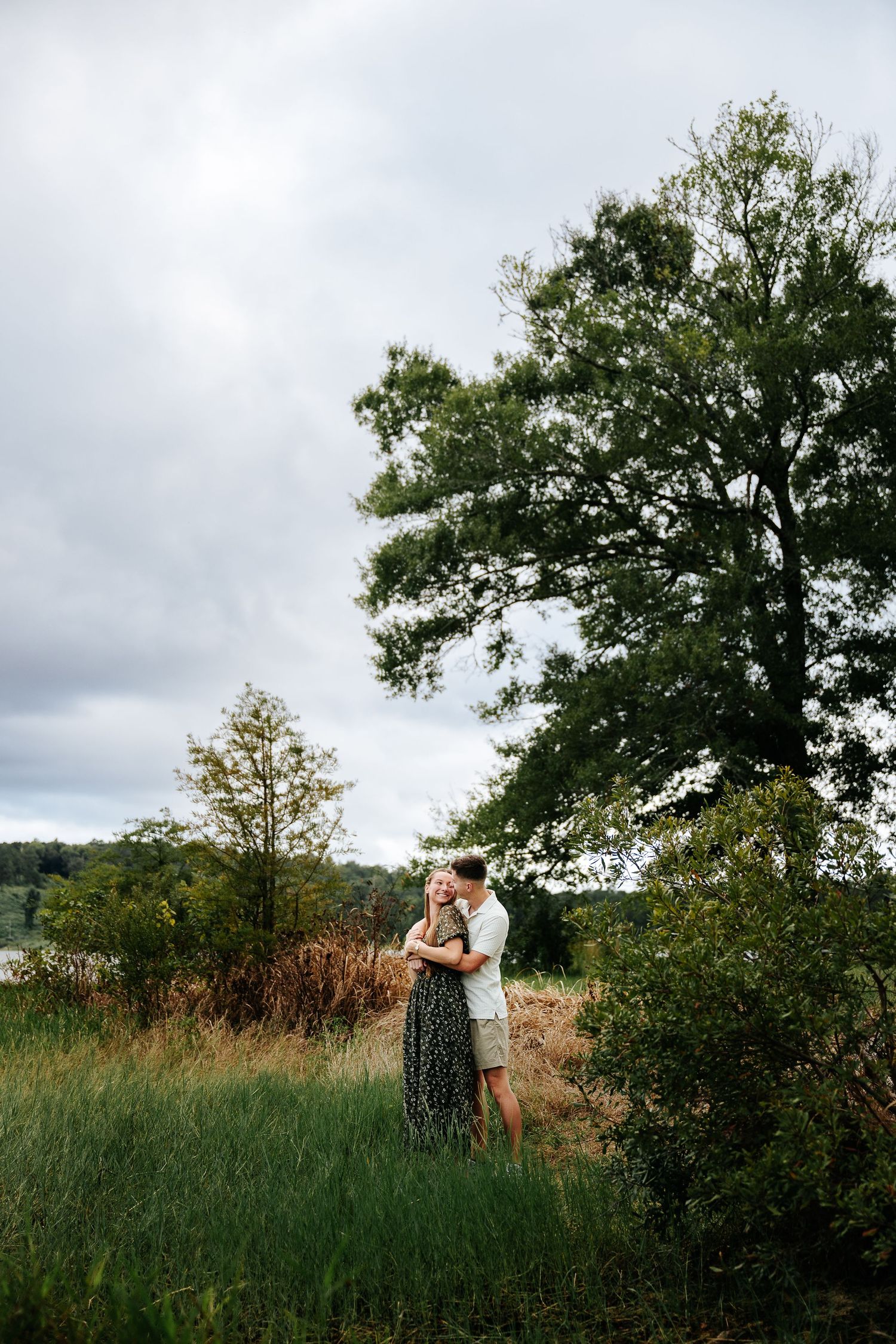 Couple embracing in a lush green field with tall trees and cloudy sky in the background.