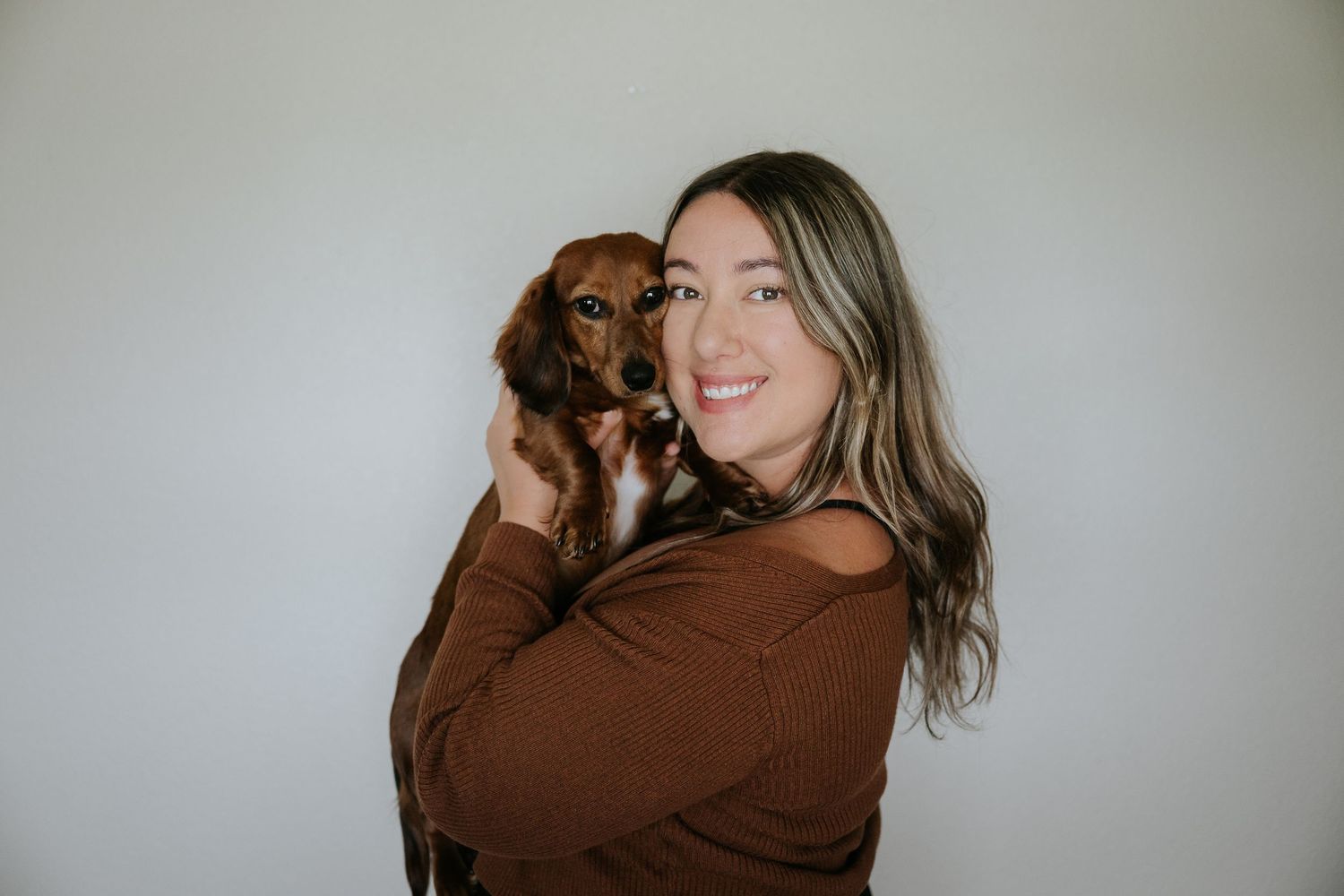Smiling woman in brown top holding a small brown dachshund puppy against a light gray background.