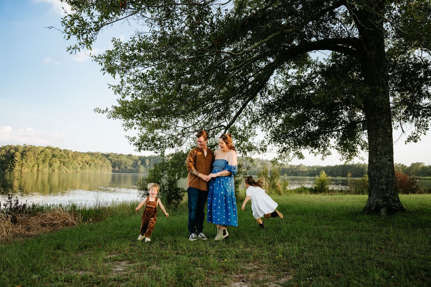 Family of four poses by a serene lake under a large tree on a sunny day in a lush green field.